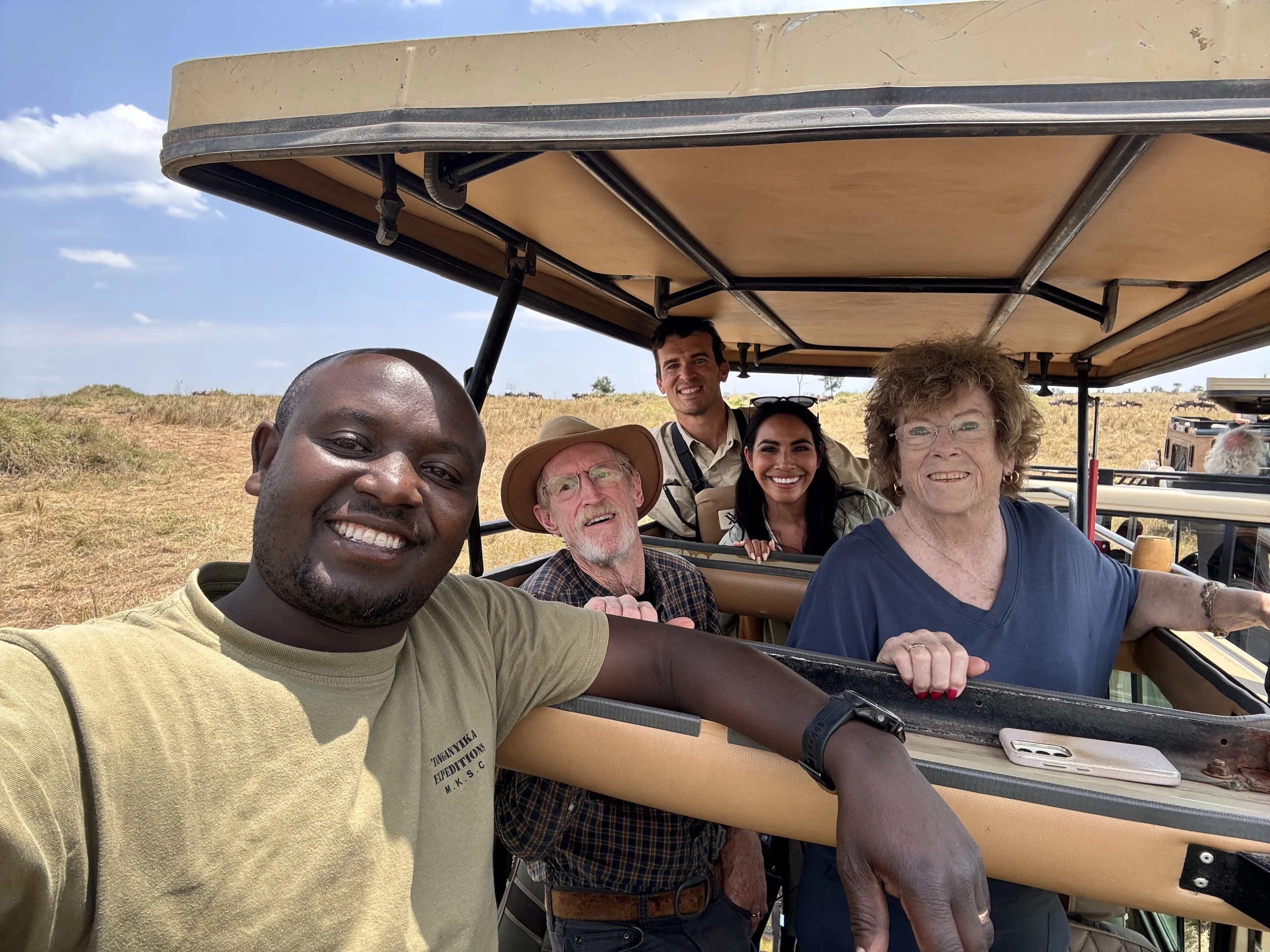 Five people on a safari adventure riding in an open-air vehicle, smiling with a dry grassland landscape in the background.