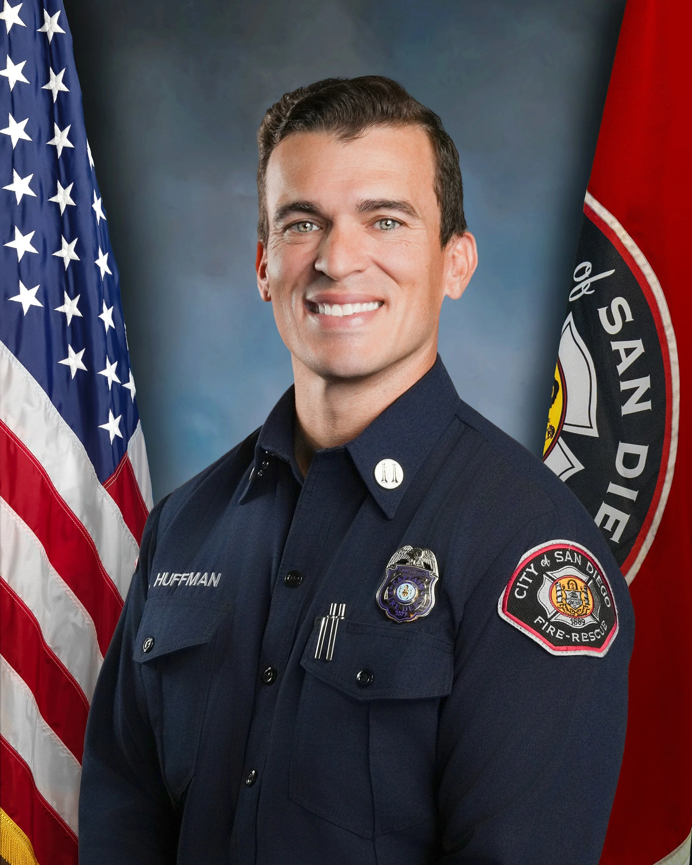 A smiling firefighter in uniform standing in front of American and San Diego fire department flags.