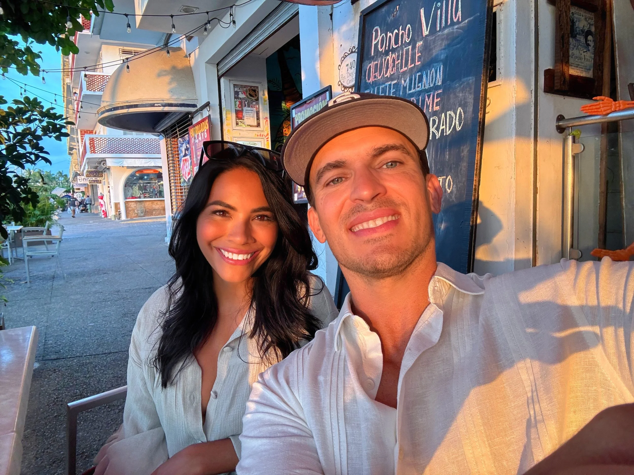 A smiling man and woman taking a selfie outside a restaurant during golden hour sunset. The woman has long dark hair and is wearing sunglasses and a light-colored shirt. The man is wearing a beige cap and a white shirt. Behind them, a chalkboard menu lists various dishes, and there are outdoor tables and chairs along a lively street with buildings and people in the background.
