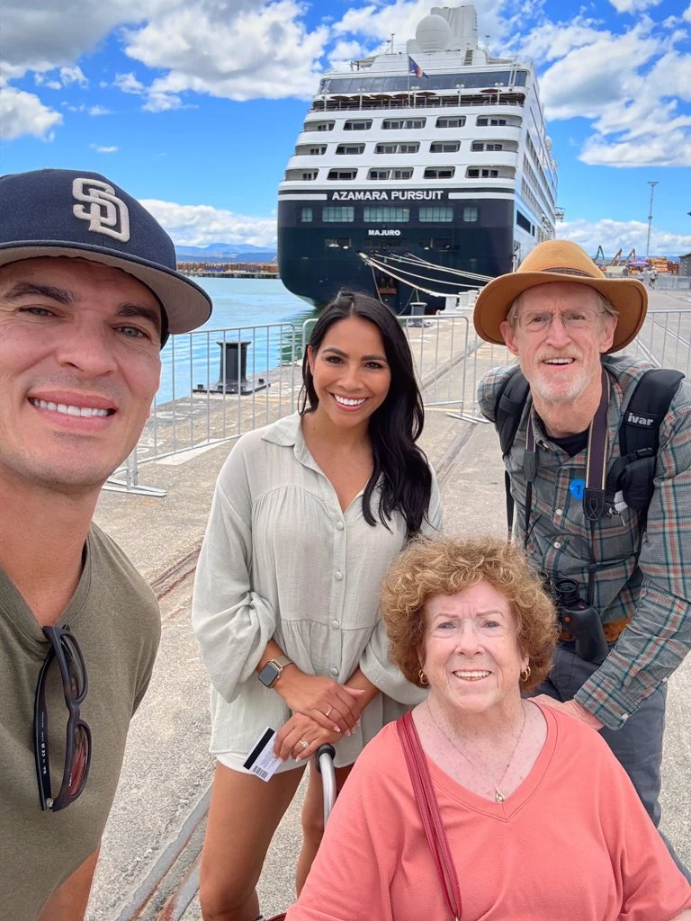 Four people taking a selfie near a dock with a large cruise ship named Azamara Pursuit in the background under a partly cloudy sky.