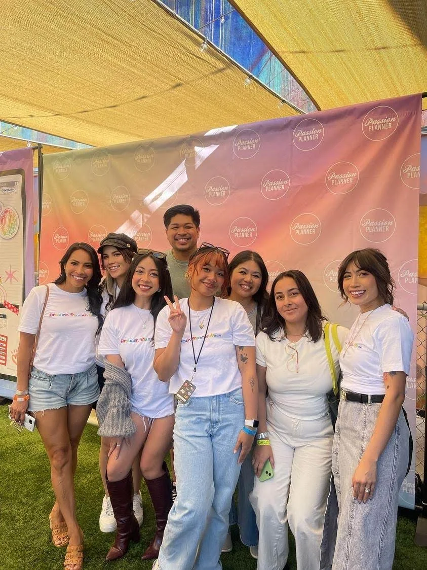 Group of seven smiling women and one man posing in front of a pink Passion Planner backdrop at what appears to be an outdoor event or festival.