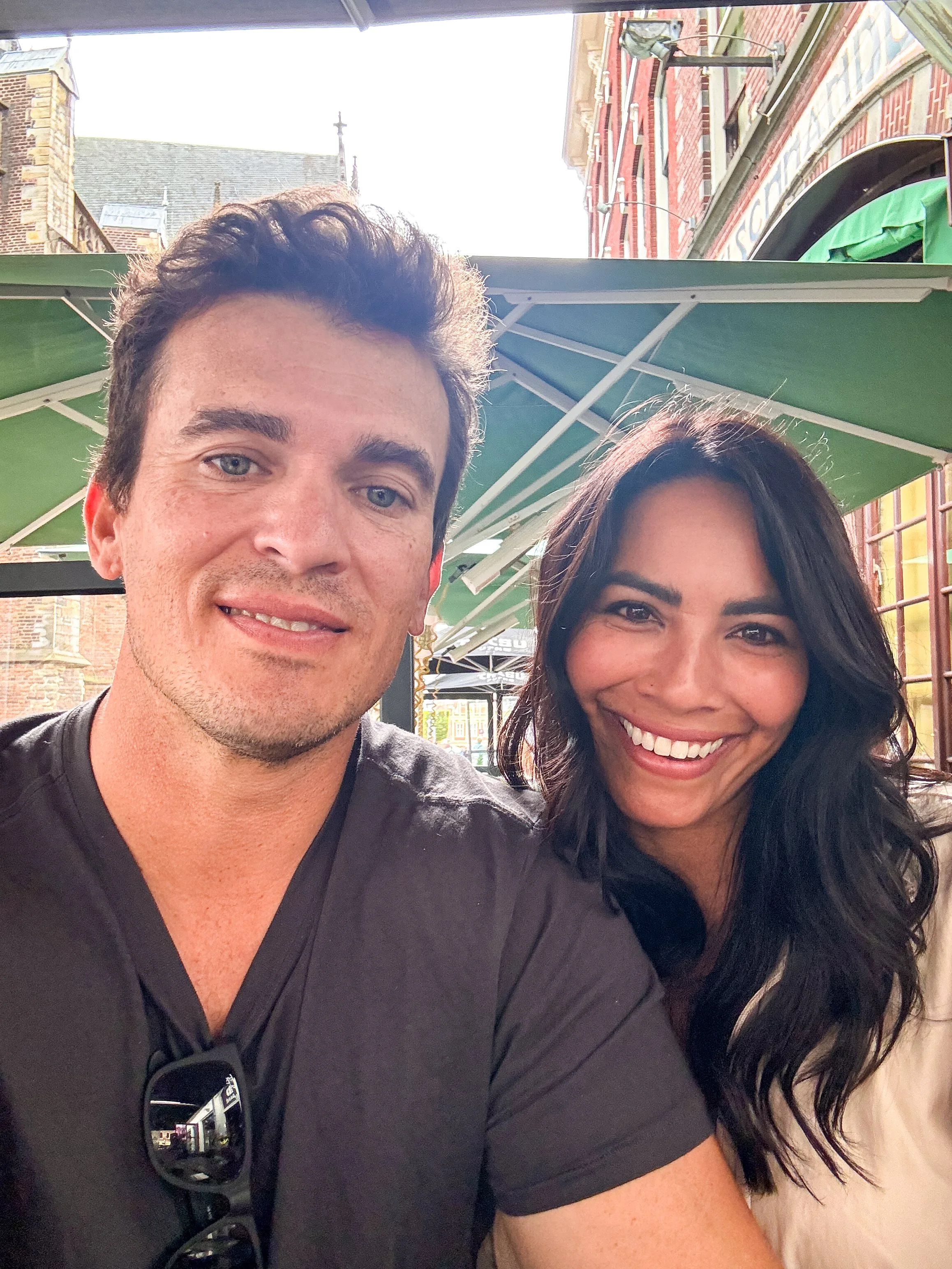 A man and woman smiling for a selfie at an outdoor café or restaurant with green umbrellas, brick buildings, and a church steeple in the background.