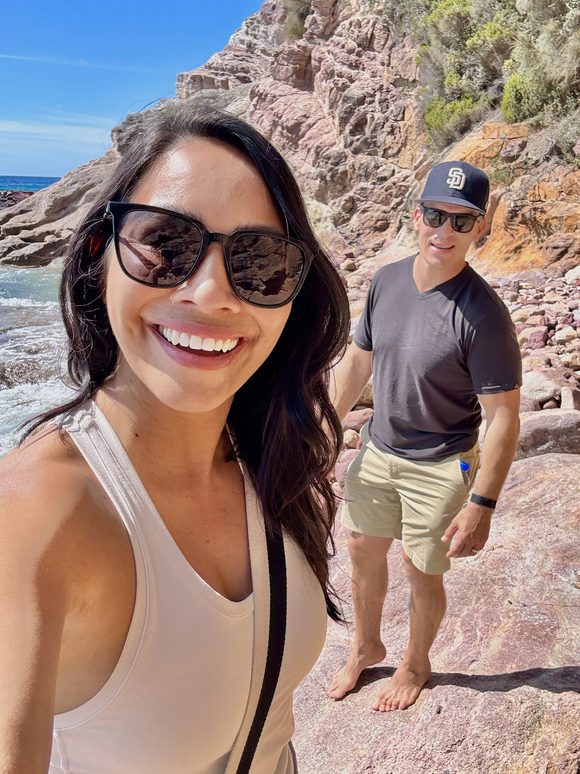 A woman with long dark hair wearing sunglasses and a light-colored tank top taking a selfie on a rocky beach with a smiling man in a black t-shirt, beige shorts, sunglasses, and a San Diego Padres cap in the background. They are near the ocean with a rocky cliff and blue sky.