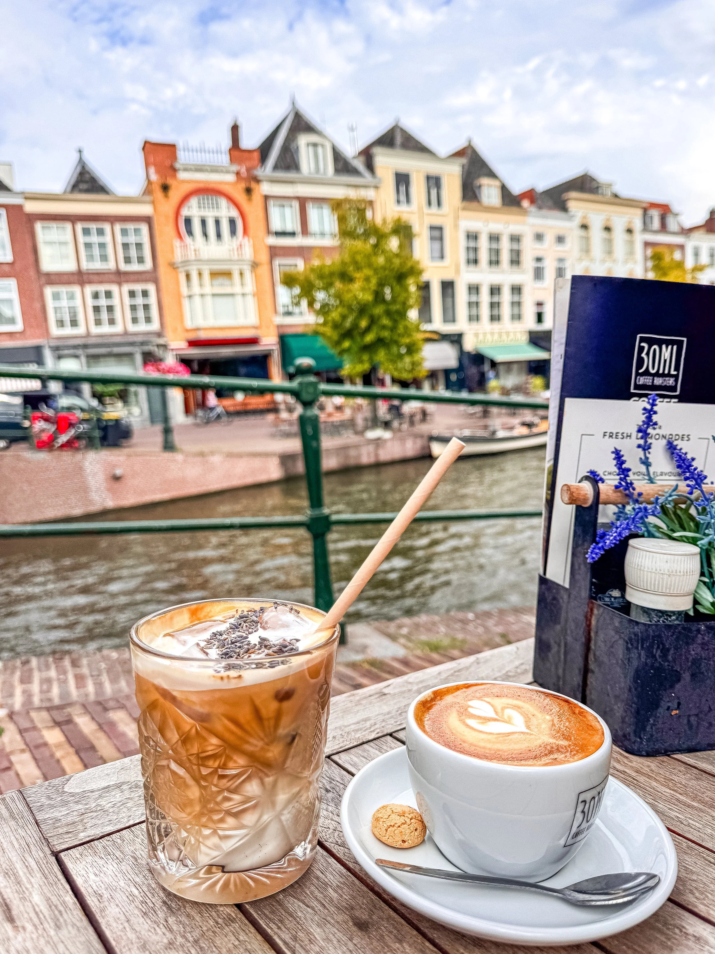 Two coffee drinks on a wooden table outside near a canal with colorful buildings in the background. One drink is iced with a straw, and the other is a hot latte with latte art, a cookie, and a spoon.