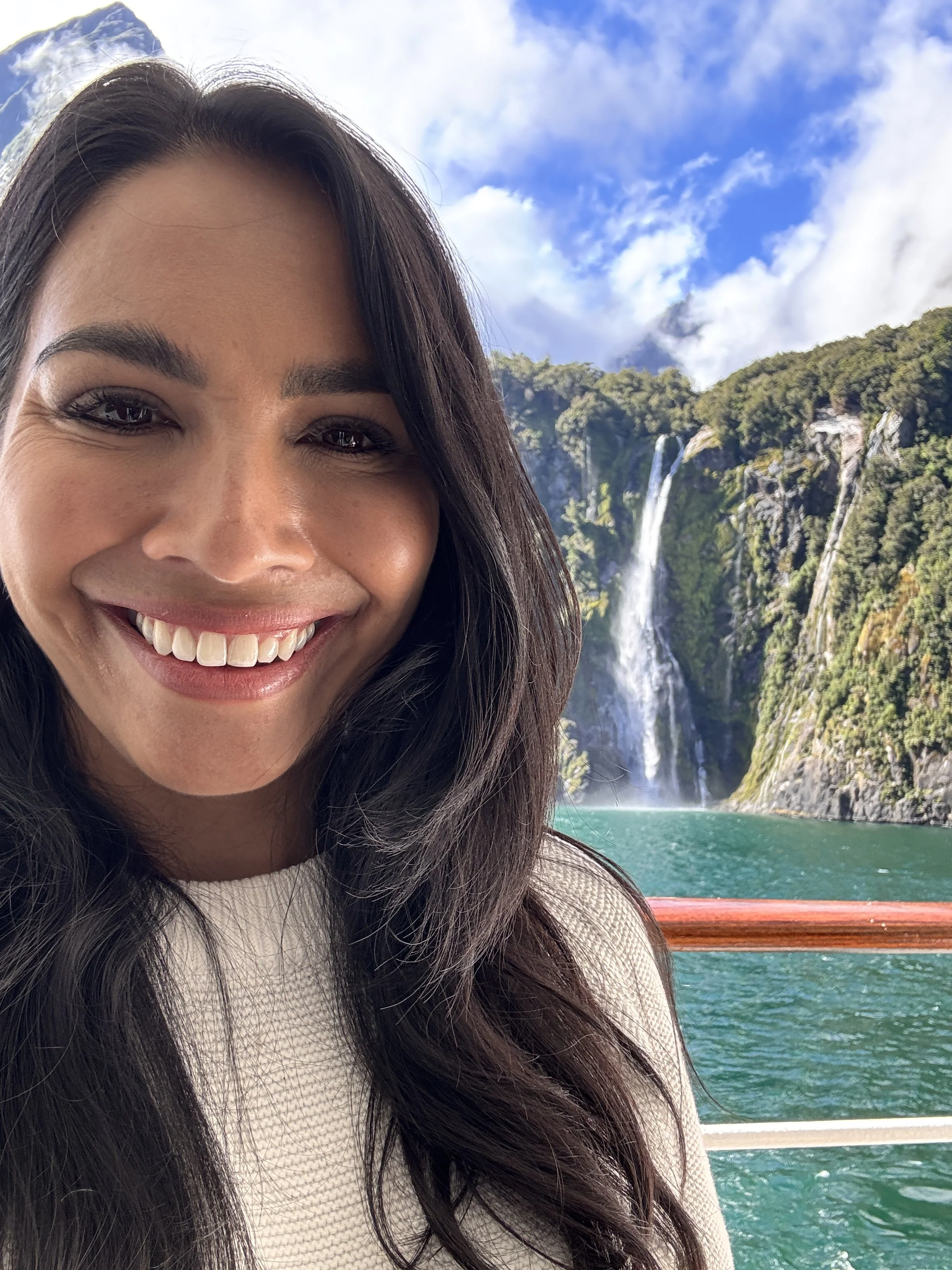 A woman smiling on a boat with a waterfall, cliffs, lush green trees, and a partly cloudy sky in the background.