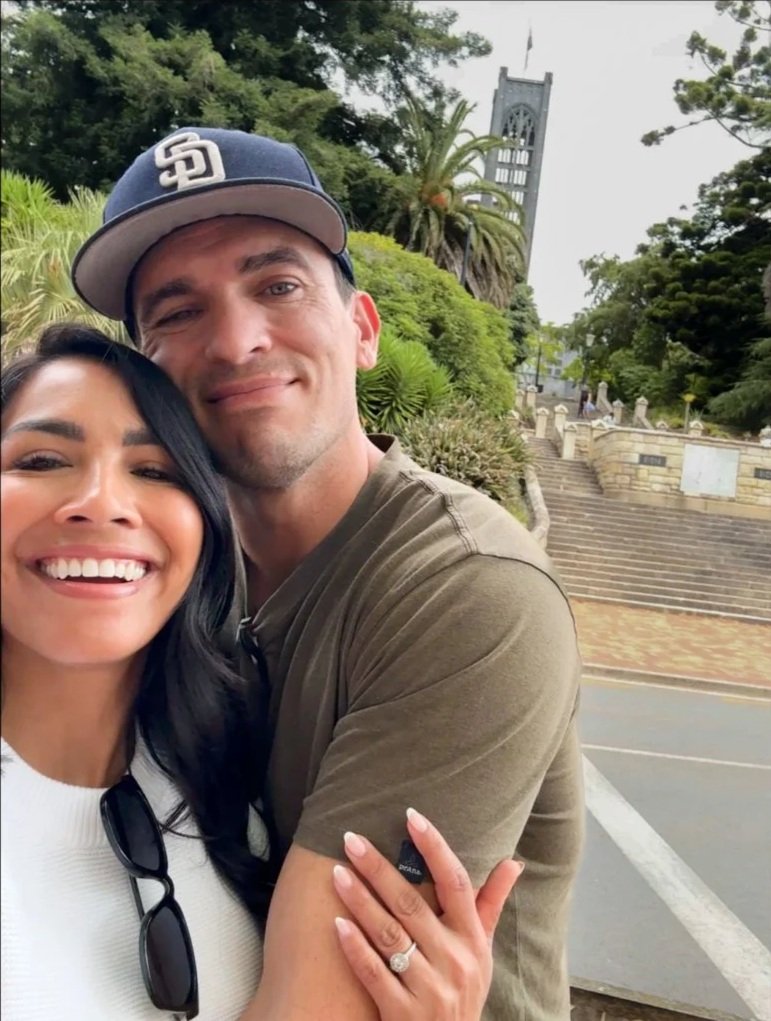 A pedestrian street with shops, outdoor seating, and potted plants under large trees, in a historic downtown area.