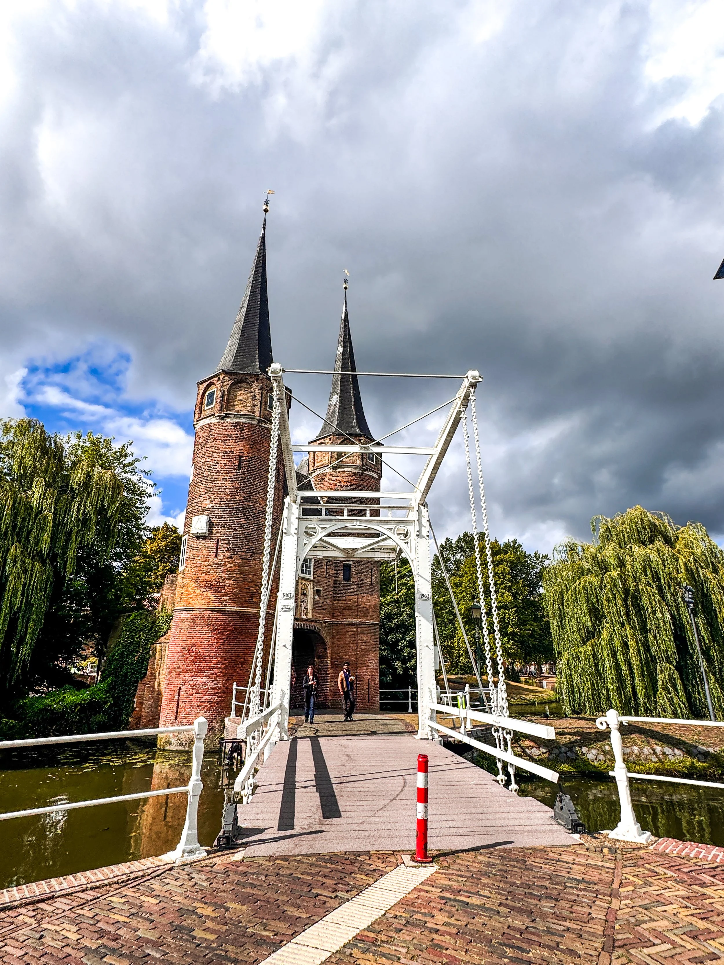Historic brick castle with pointed towers behind a white swing bridge over a small waterway, surrounded by trees with cloudy sky overhead.