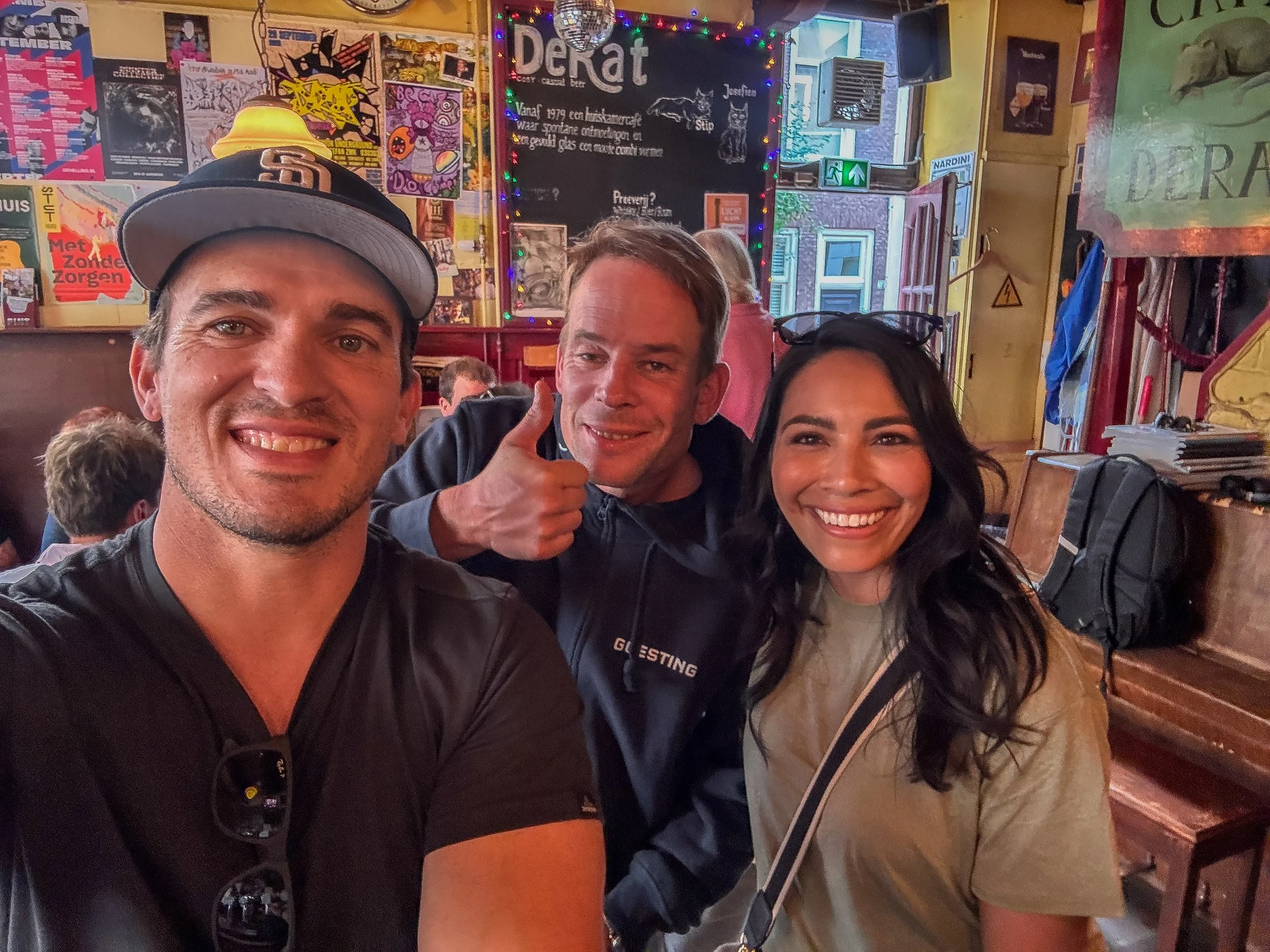 Three friends taking a selfie inside a lively bar, surrounded by colorful posters, bright lights, and eclectic decorations.