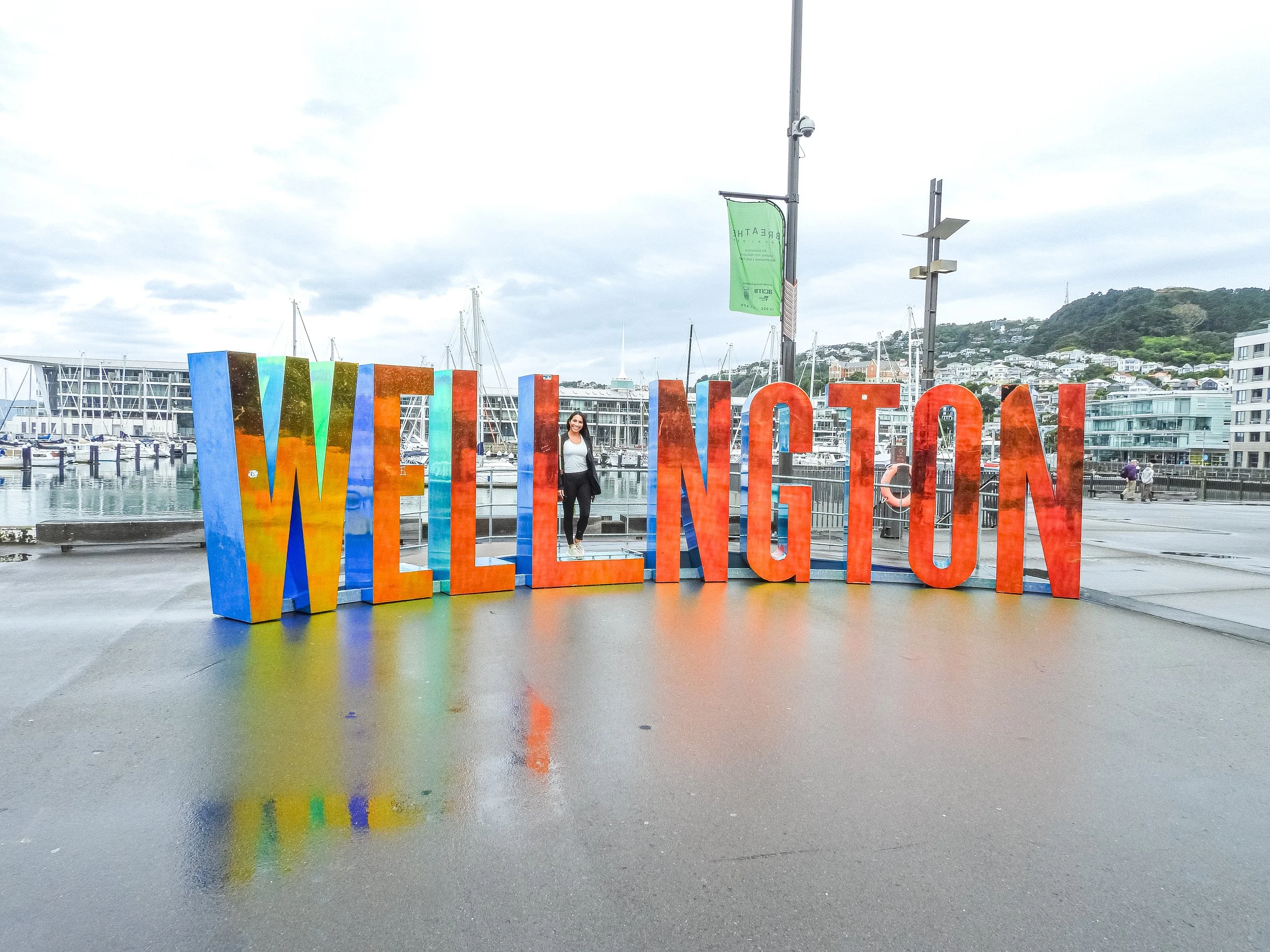 Colorful large letters spell 'WELLINGTON' at a waterfront with boats, a dock, and buildings in the background, under an overcast sky.