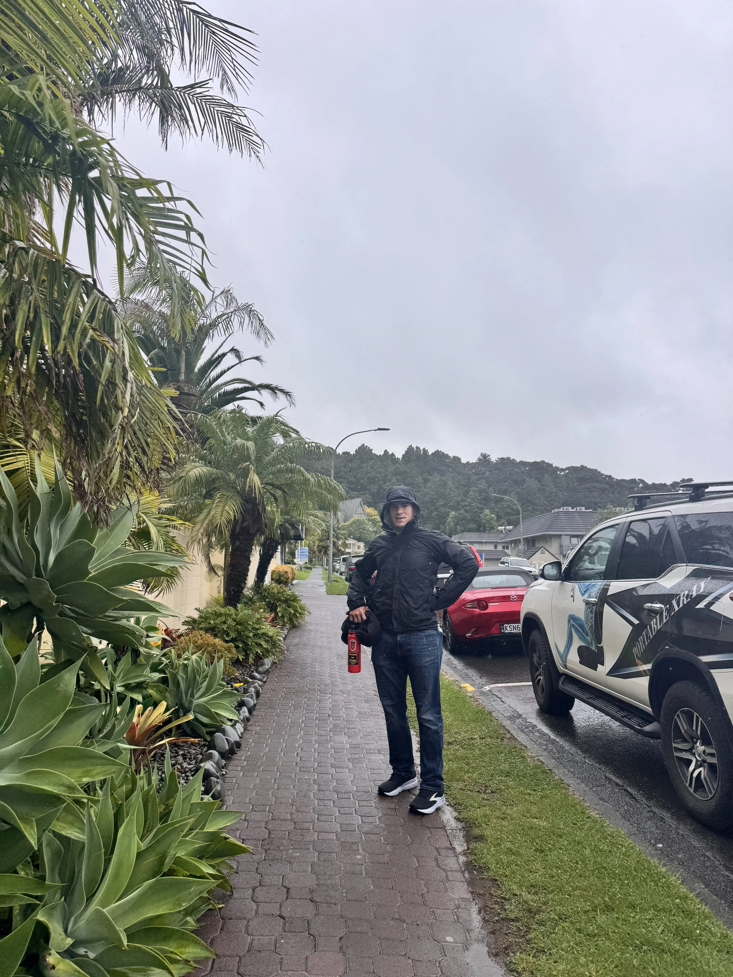 Man standing on a rainy sidewalk near parked cars and lush greenery, holding a fire extinguisher, wearing a black jacket and jeans.