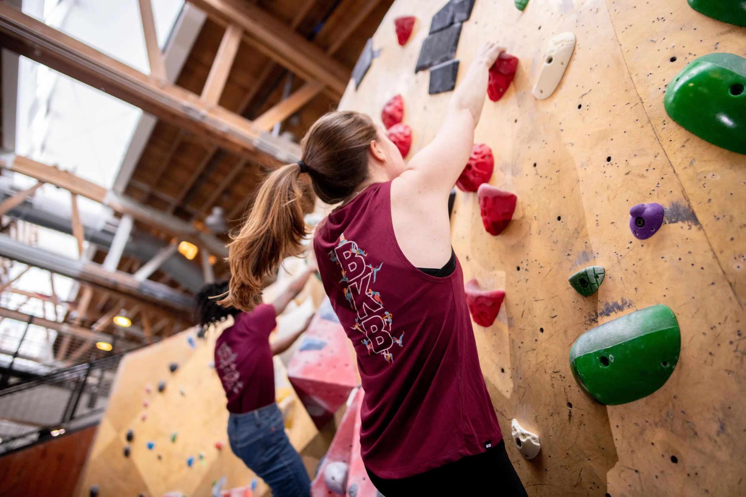 Two people wearing maroon shirts climbing an indoor bouldering wall with colorful holds.