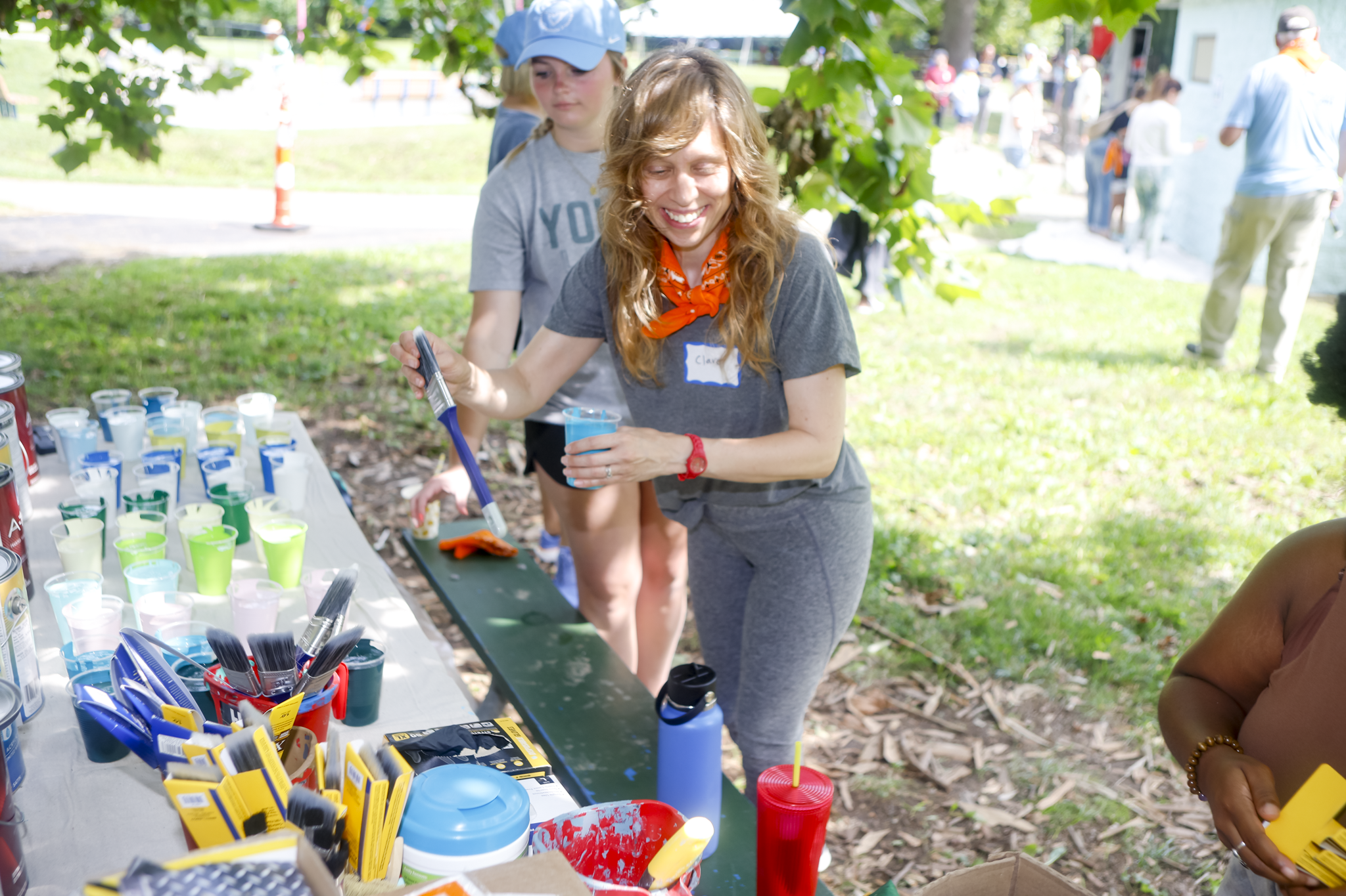 A woman smiling while filling a cup with blue liquid at an outdoor table with colorful cups and supplies, with children and people in the background at a park or paint-by-number event.