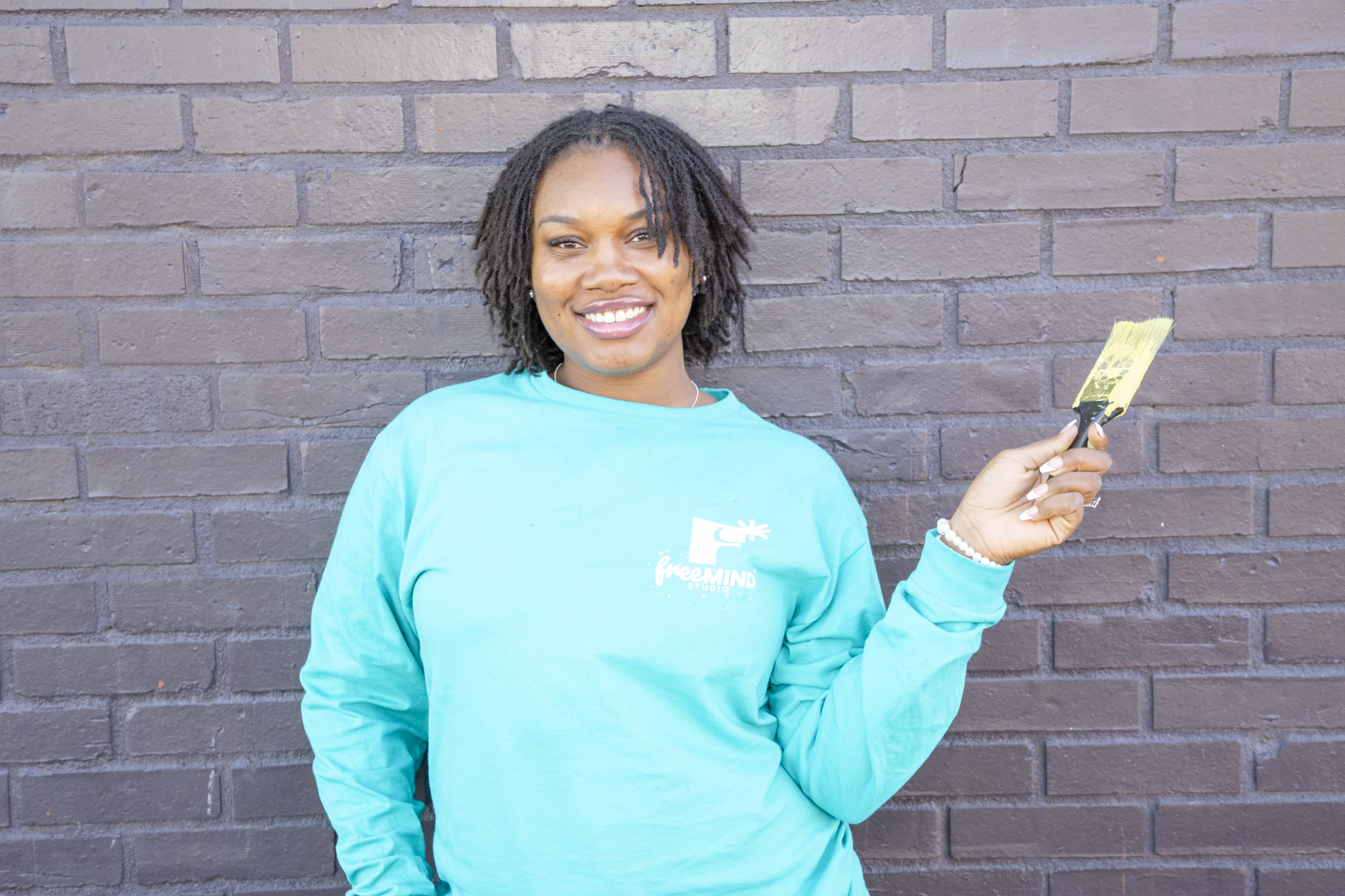 A woman with dark hair styled in dreadlocks smiling, wearing a turquoise long-sleeve shirt with a logo that reads 'freeMIND' and holding a paintbrush in her right hand, standing against a brick wall.