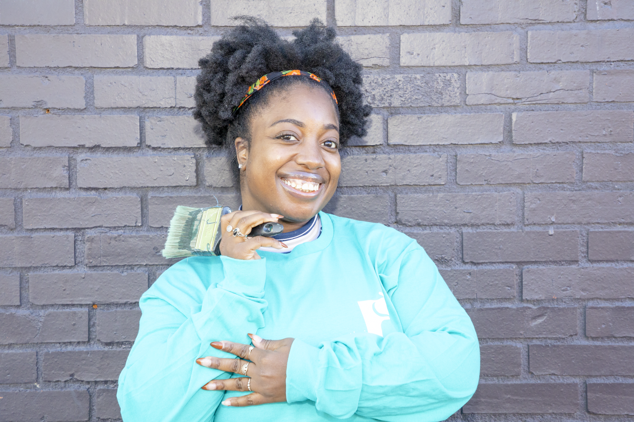 A woman with dark, curly hair in a bun, wearing a headband and a light blue sweatshirt, standing in front of a brick wall. She is smiling and holding a paintbrush near her face.