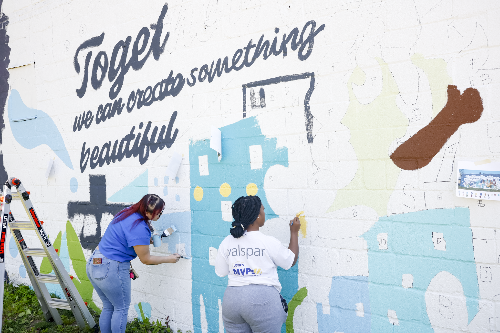 Two women painting a paint-by-number mural on a white brick wall, with the words "Together we can create something beautiful" written in black cursive at the top. One woman is painting with blue paint, and the other is painting with yellow paint.