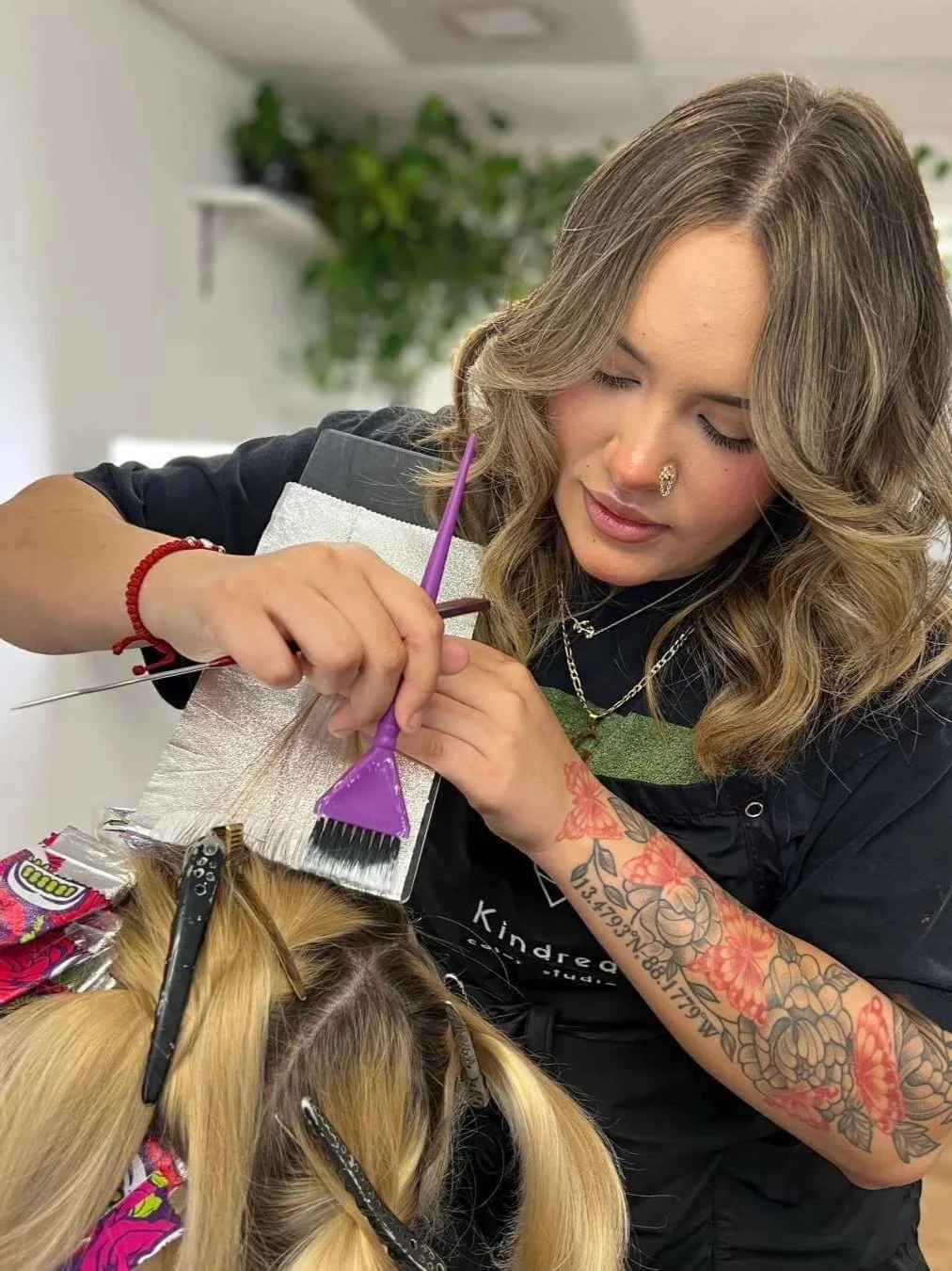 Gaby at Kindred Color Studio  working on a client's blonde hair in a salon, holding a small comb and a hair color application tool, with hair clips securing the client's hair.