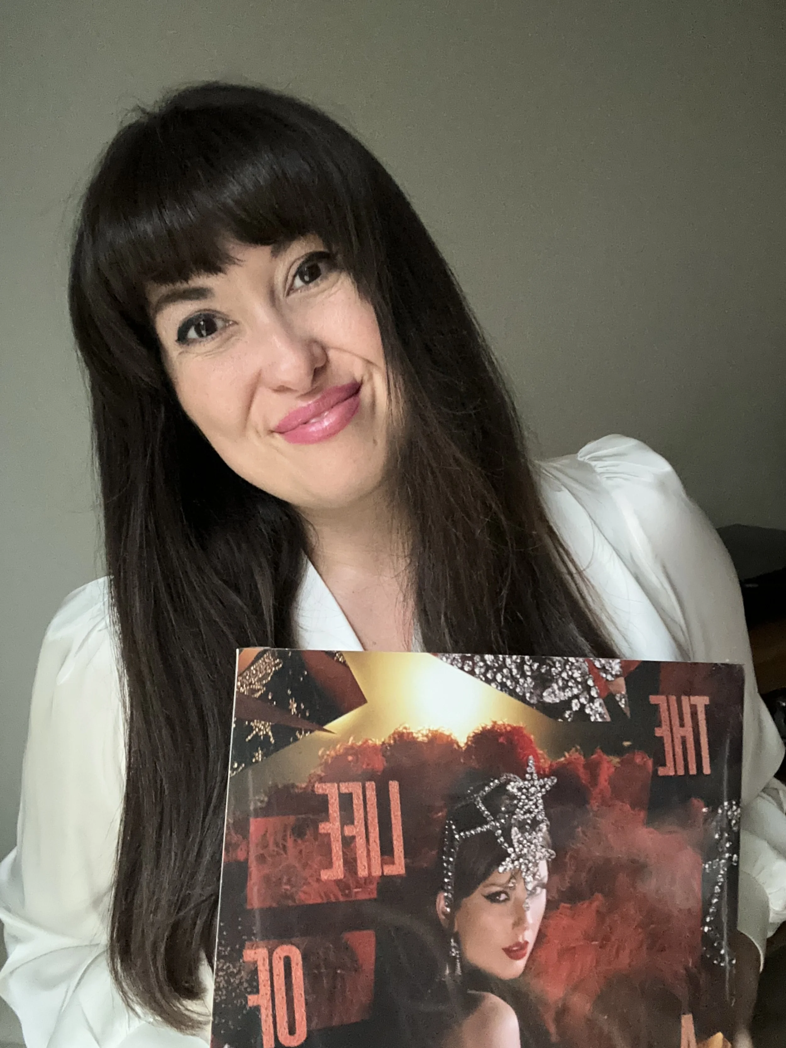 A woman with long dark hair and freckles is smiling at the camera while holding a magazine cover featuring a woman wearing jewelry and a black dress with a fiery red background.