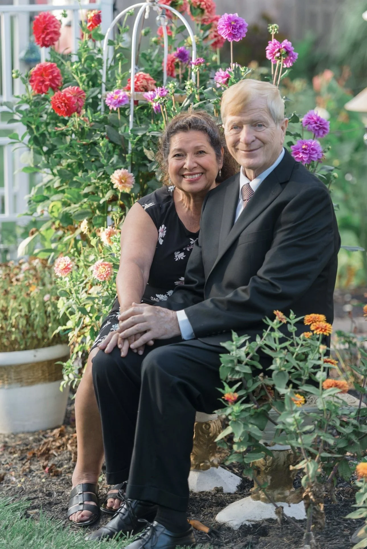 A smiling older woman in a black floral dress and a smiling older man in a black suit sit together on a garden bench, surrounded by colorful blooming flowers.