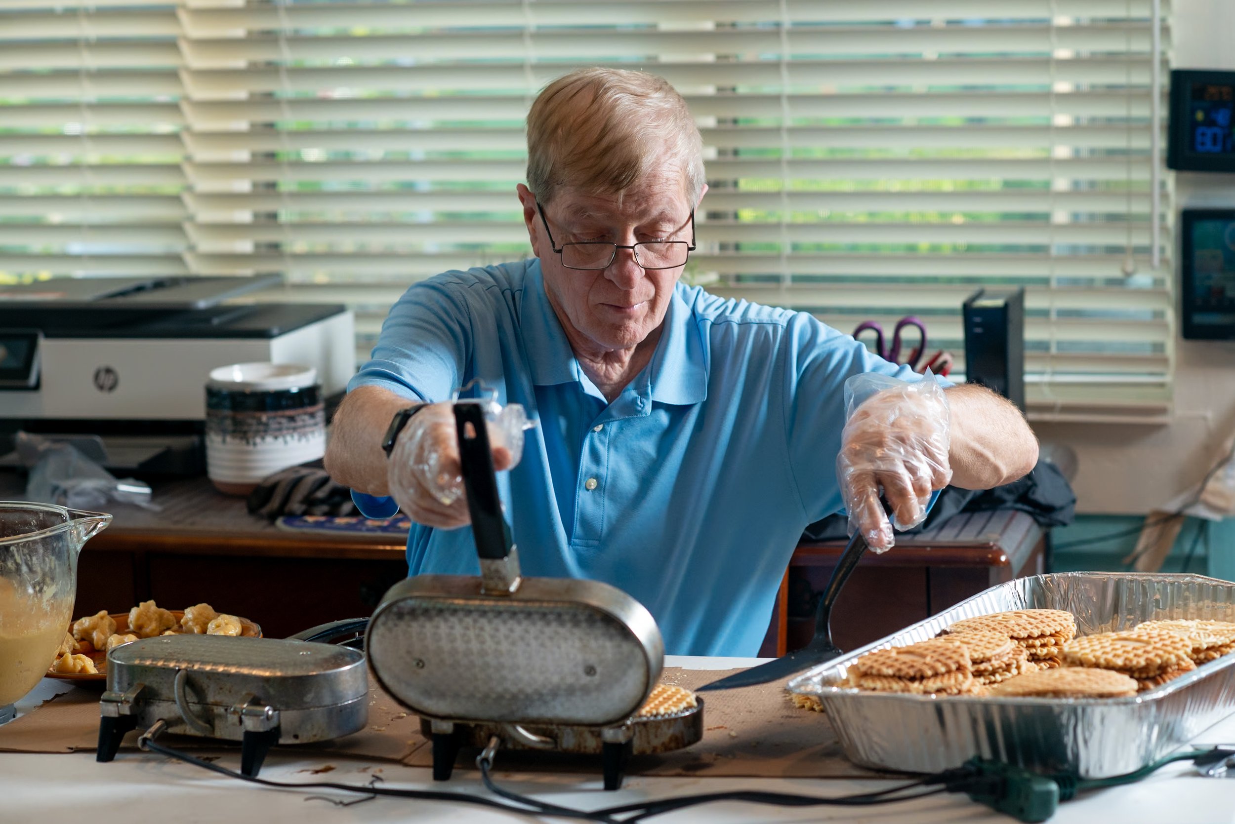 An elderly man in a blue polo shirt and glasses making waffles using a waffle iron, with a tray of finished waffles nearby.