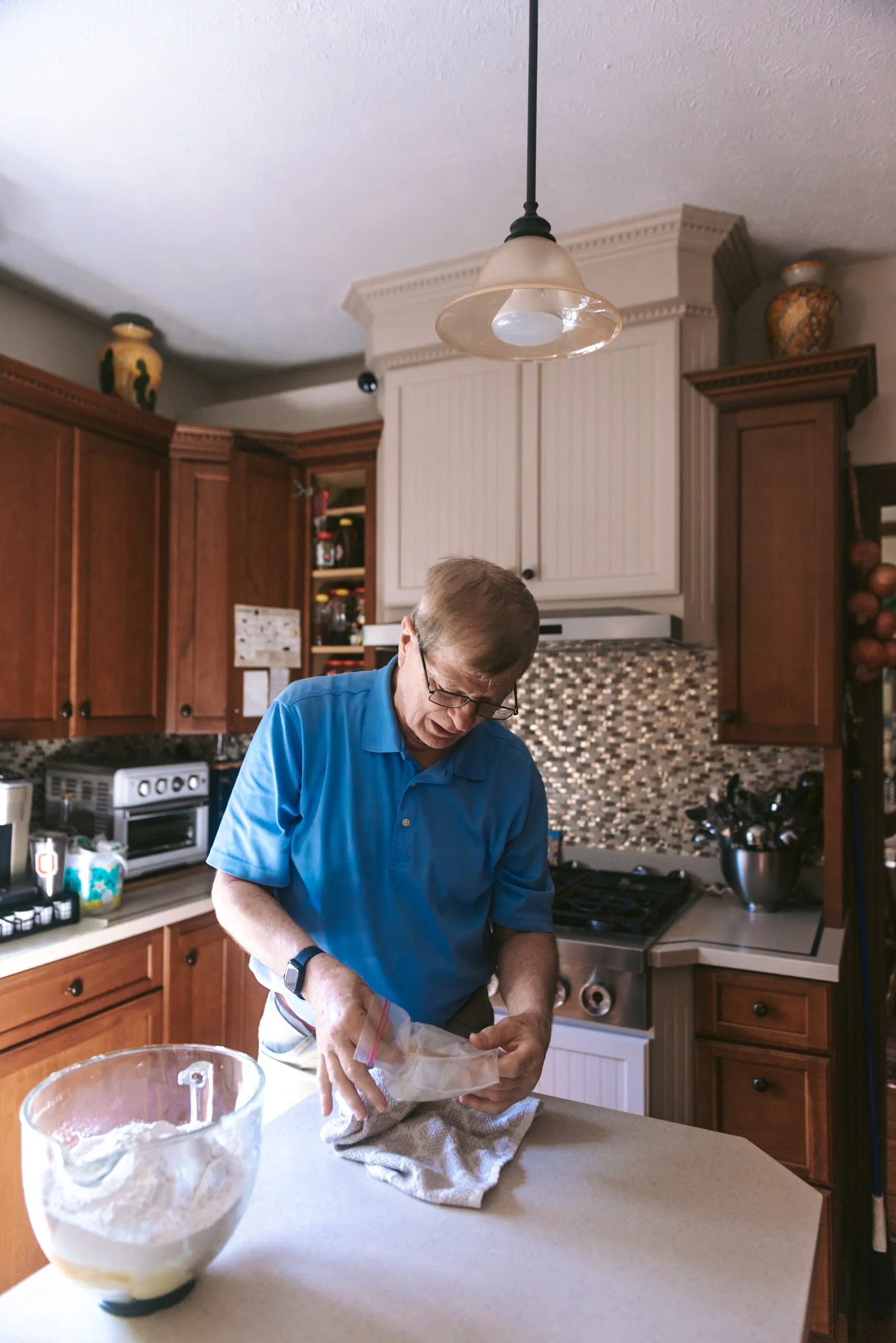 An elderly man wearing glasses and a blue shirt is in a kitchen, opening a plastic bag while standing at a kitchen island. There is a glass mixing bowl with flour on the counter in front of him, and the kitchen has wooden cabinets, a stove, and a tiled backsplash.