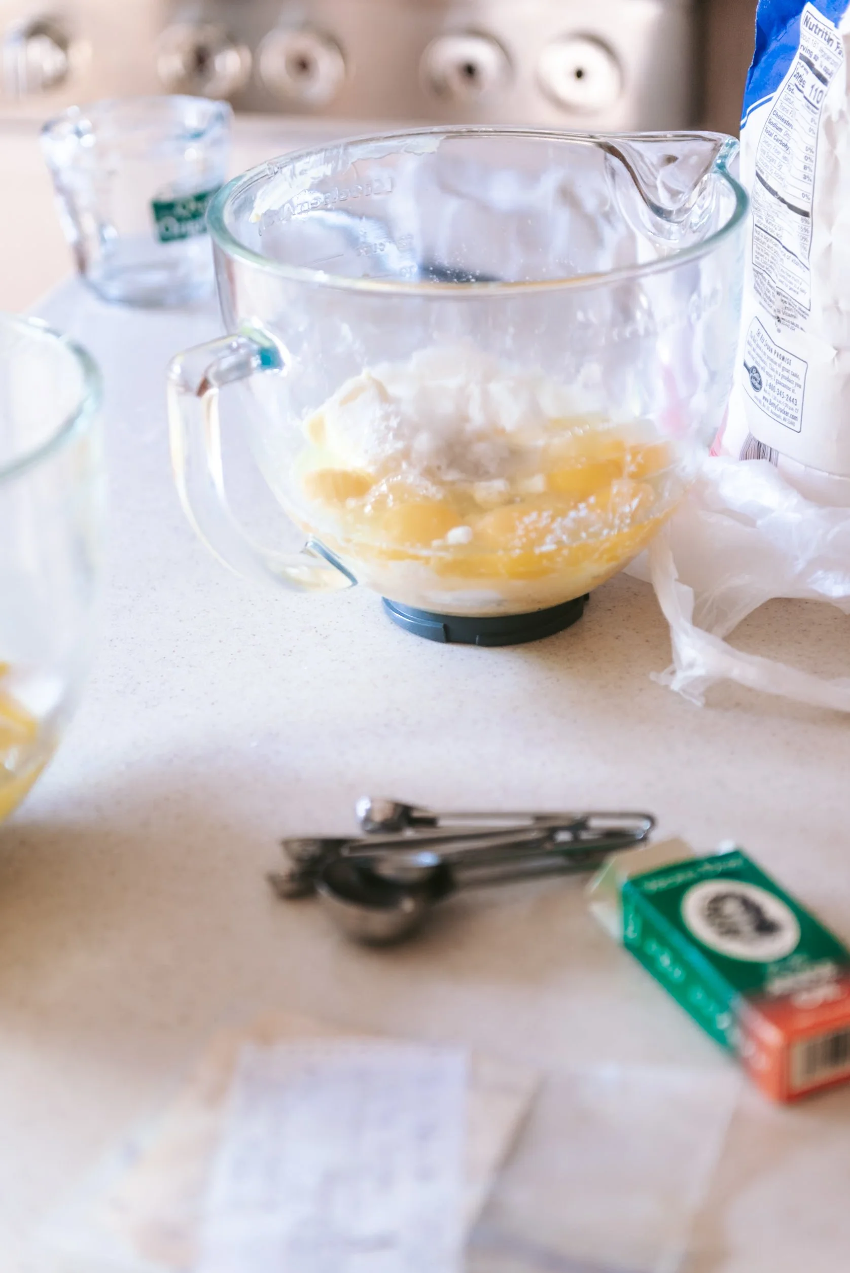 A glass mixing bowl with butter and sugar cream, on a kitchen countertop, with measuring spoons, a green box, and a package of flour nearby.