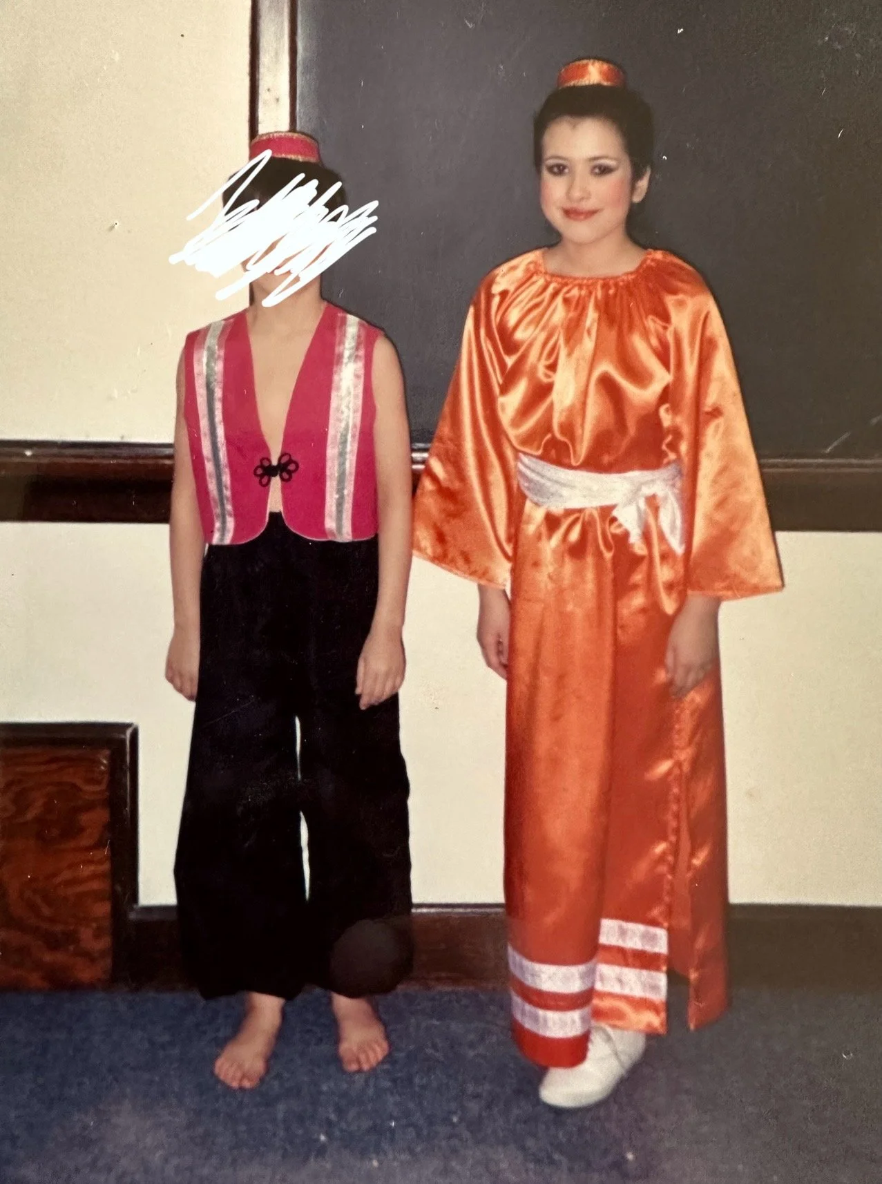 A woman in traditional Vietnamese áo dài and a girl in a pink and black dress, standing indoors in front of a dark wooden wall, with one person's face scribbled out for privacy.