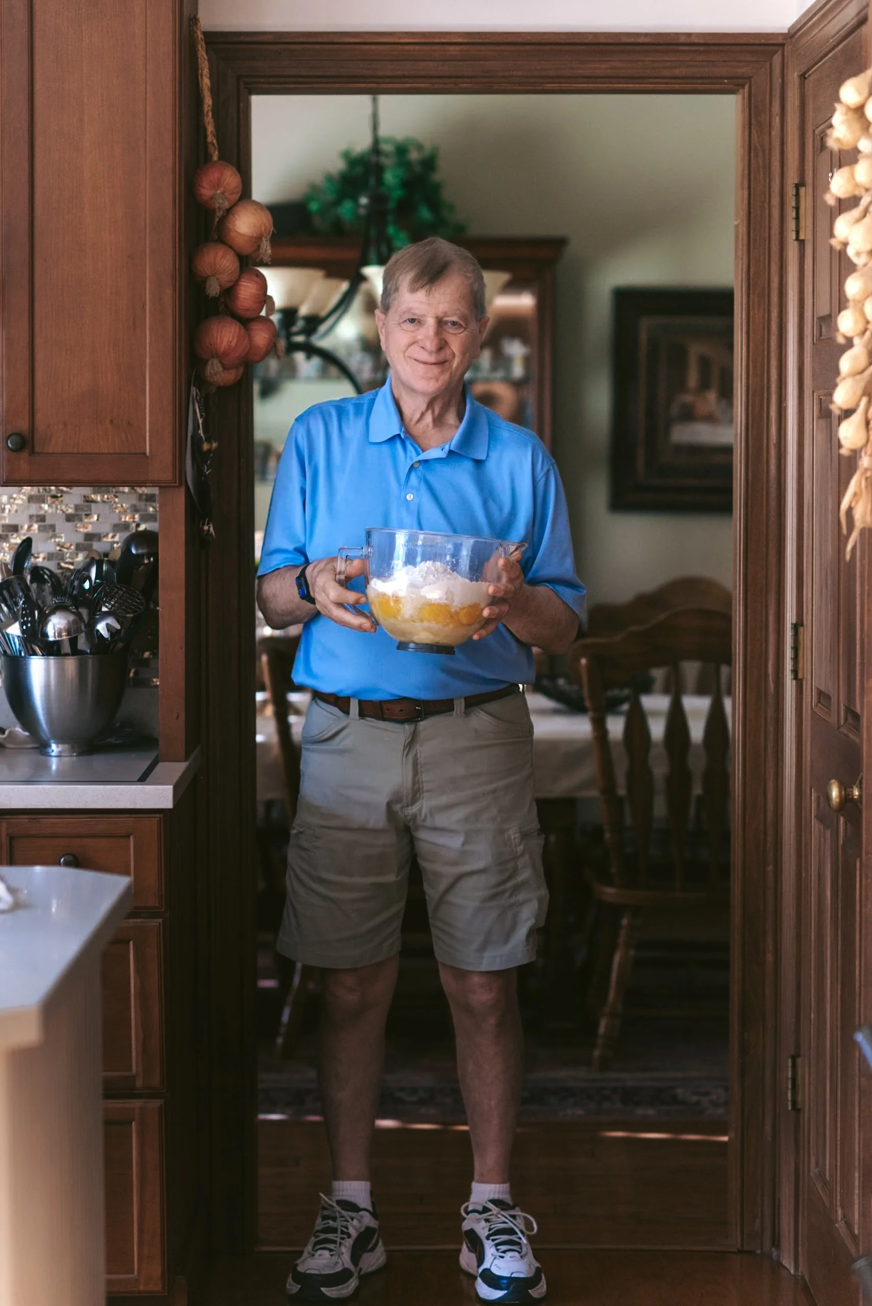 An elderly man in a blue polo shirt and beige shorts stands in a doorway, holding a mixing bowl with ingredients. There are kitchen cabinets and a dining area in the background.