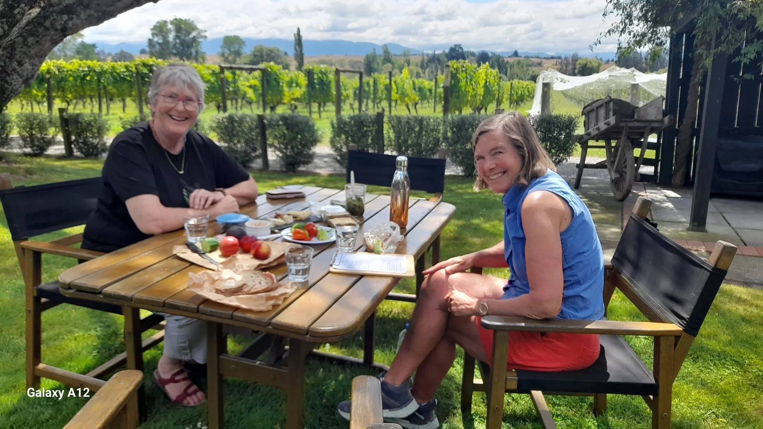 Two women sit at a wooden outdoor dining table with food and drinks, smiling, with a vineyard and mountains in the background.