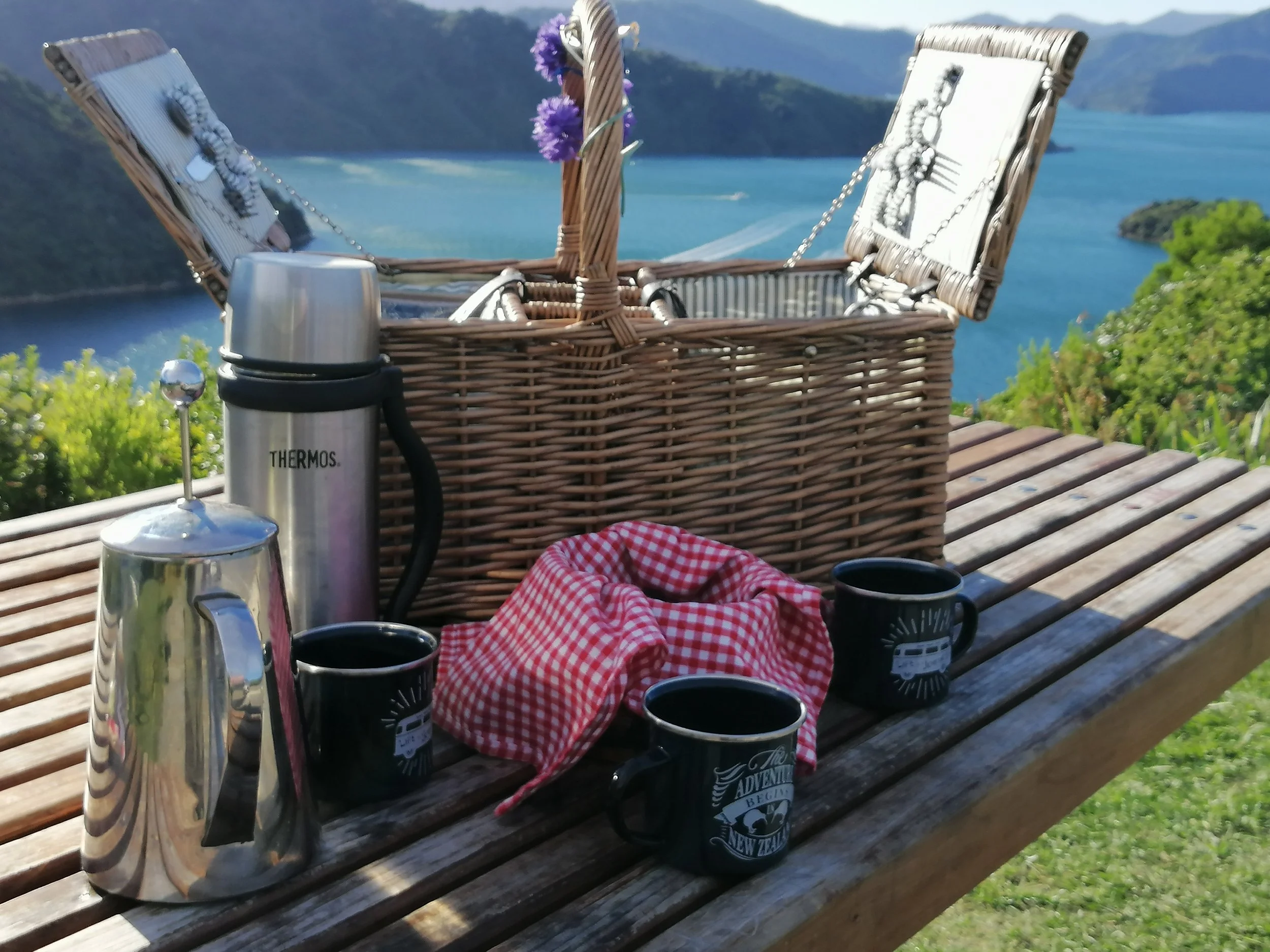 A picnic table outdoors with a black mug, a thermos, and a red and white checkered cloth. A wicker basket with purple flowers and a silver kettle is present, overlooking a scenic view of water and hills in the background.