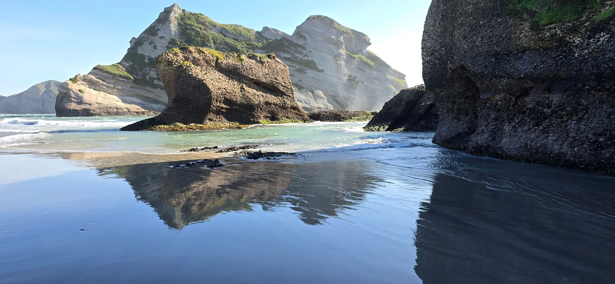 A scenic coastal landscape at Wharariki Beach with large rocky cliffs towering over the ocean, sandy beach, and calm water reflecting the rocks and sky.