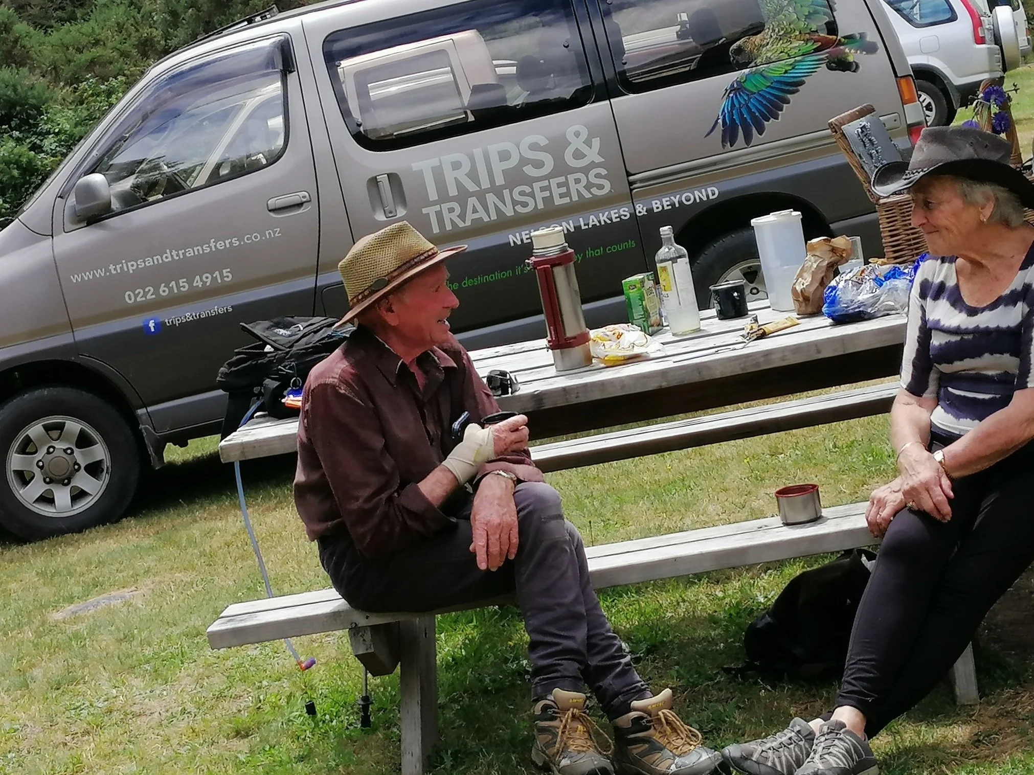 A woman and an elderly man are sitting on a park bench outdoors, having a conversation with a smile. There is a table with snacks and drinks between them, and a gray van with the label 'Trips & Transfers' and a colorful bird decal parked nearby.