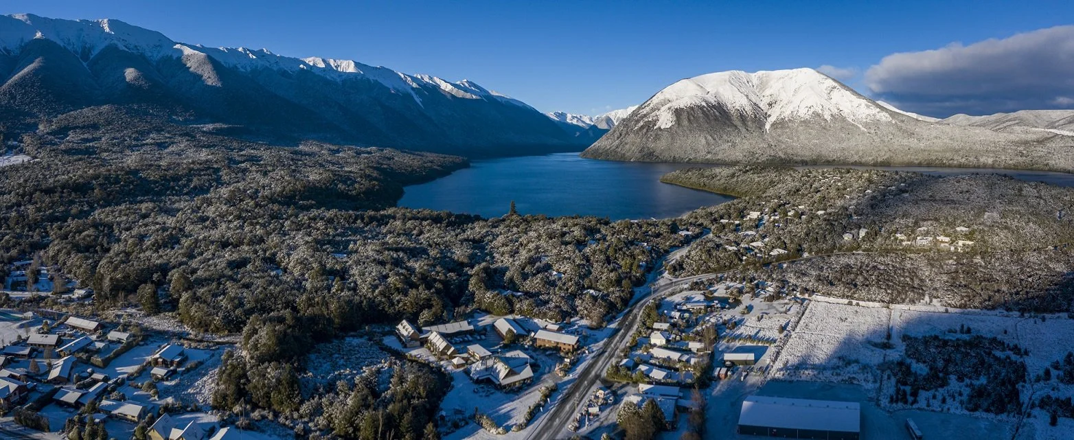 A snow-covered village with houses along a winding road, surrounded by dense forest, with a large lake and mountains in the background under a blue sky with some clouds.