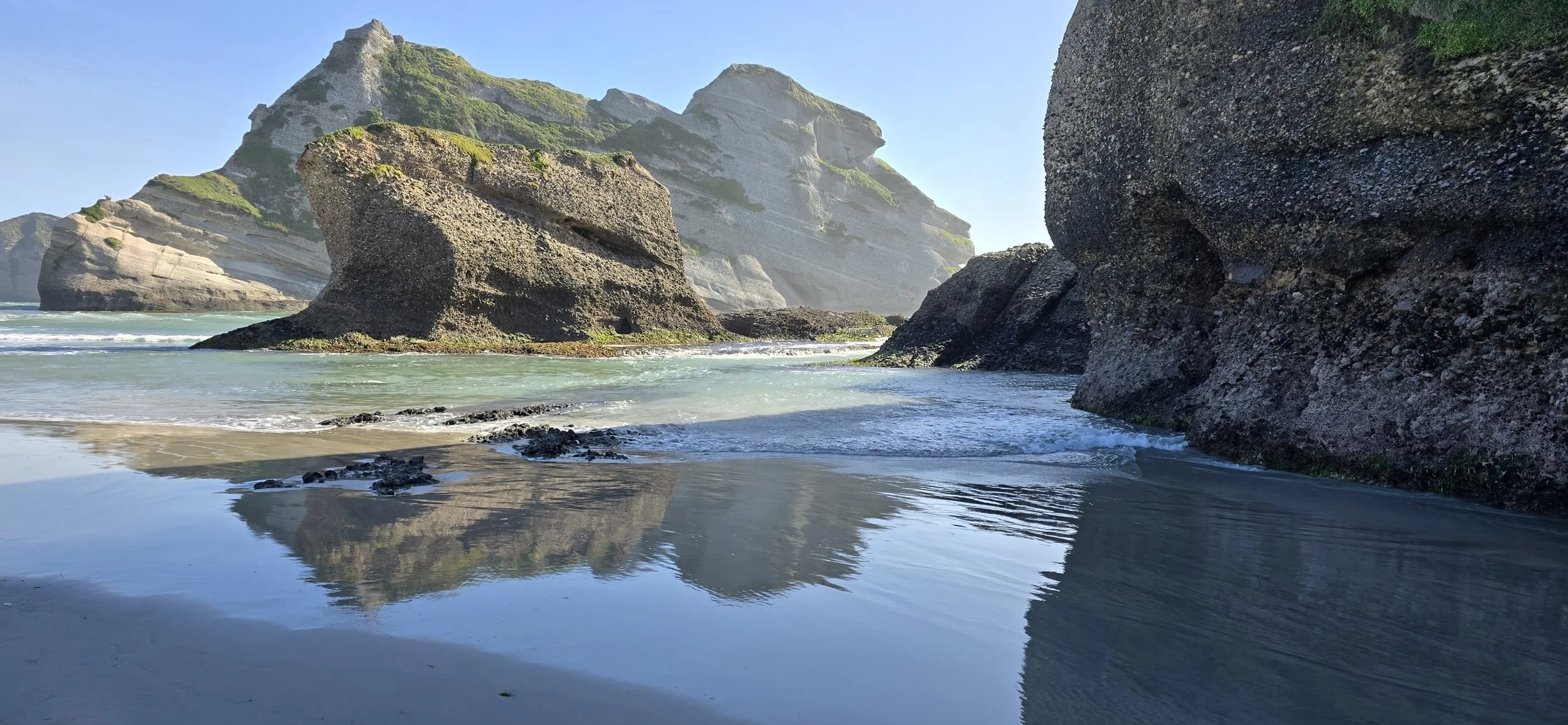 Rock formations and cliffs along a sandy beach at Wharariki Beach with calm ocean water and blue sky.