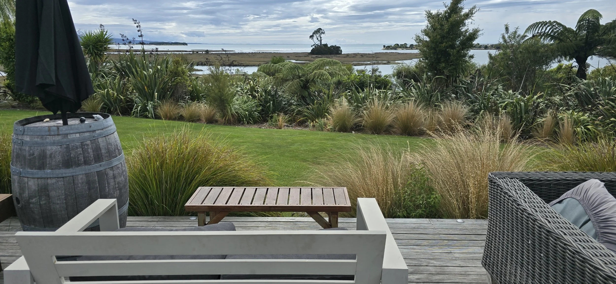 A scenic outdoor view from a deck at Zatori Lodge in Collingwood, showing a grassy yard with shrubs and trees, overlooking a body of water with small islands and trees in the distance, under a cloudy sky.