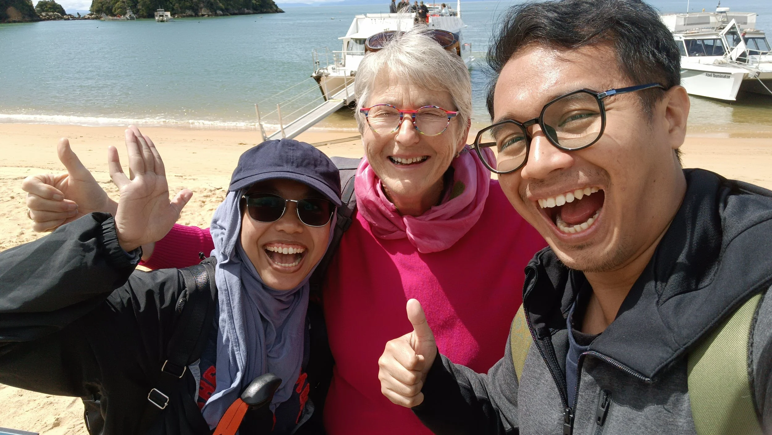 Three smiling people taking a selfie on a beach at kaiteriteri with boats and water in the background. They are making excited gestures with their hands, wearing sunglasses, and appear to be enjoying a sunny day.