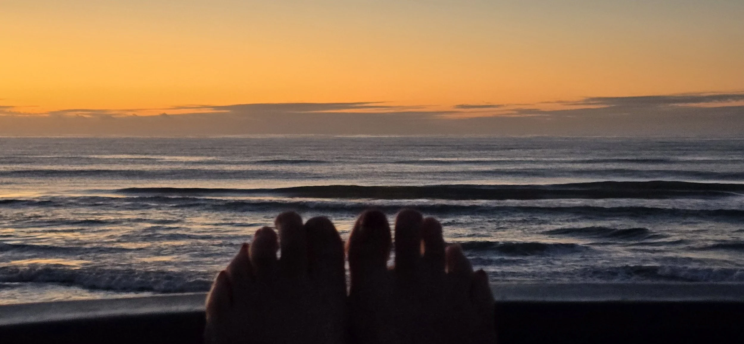 Person in a spa pool, silhouetted against a sunset over the ocean with waves.
