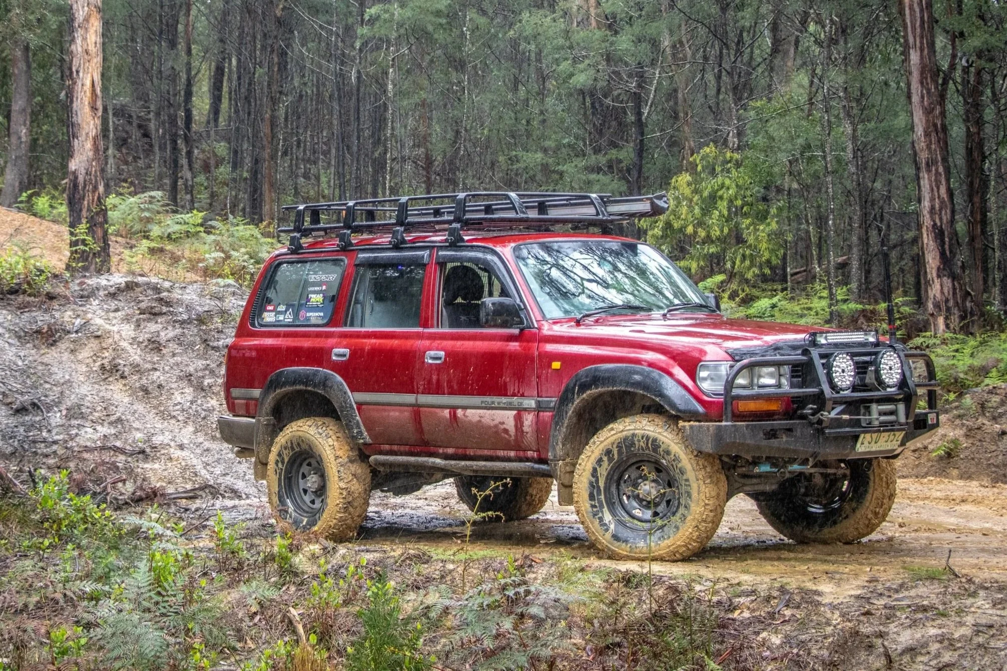 A red off-road SUV driving through a dirt trail in the Aussie bush, fitted with Windforce A/T M/T tyres with mud splattered on the tires and body. The vehicle has a roof rack and front bumper lights, and is surrounded by tall trees and green foliage.