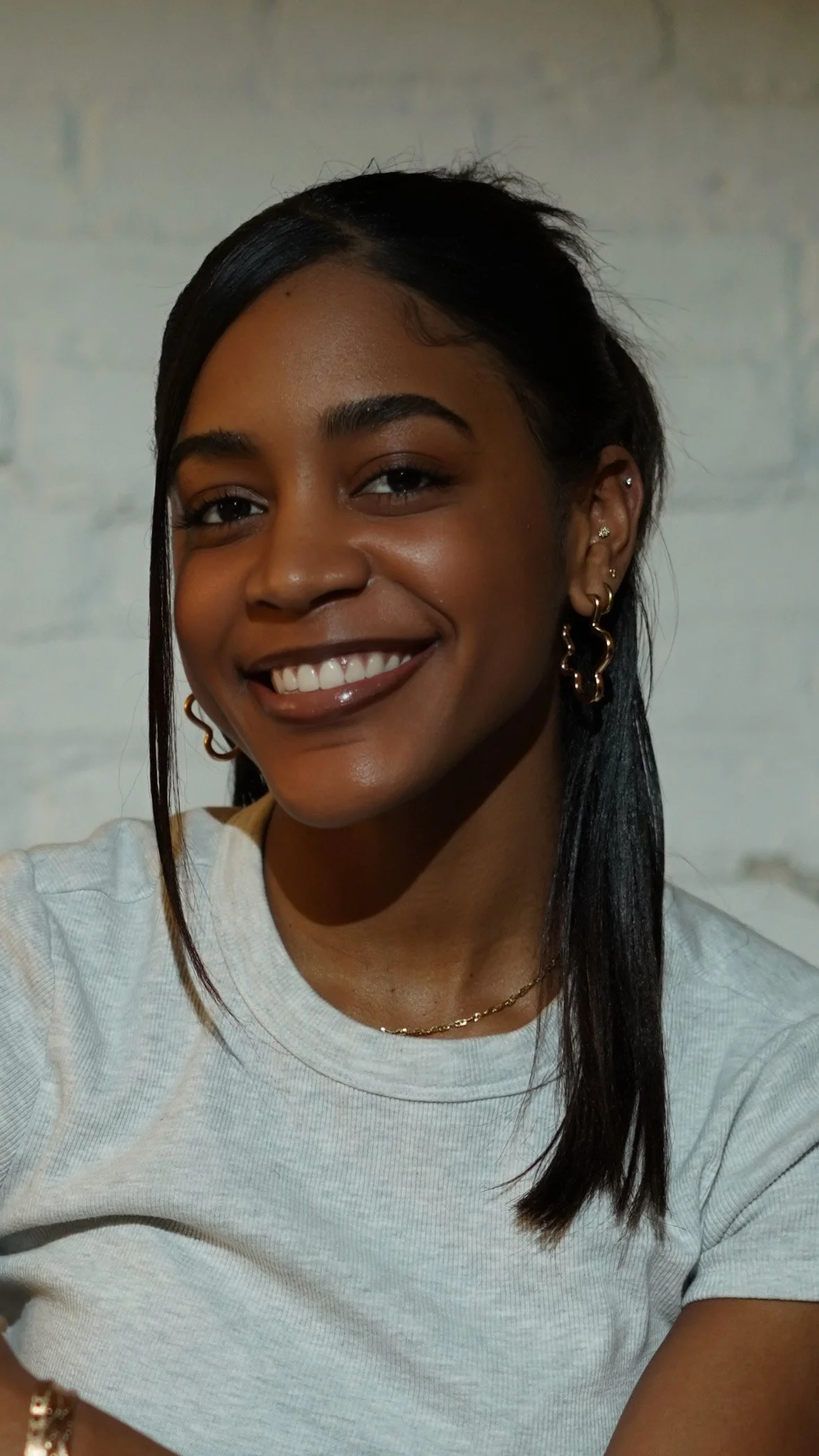 A young woman with dark hair, smiling, wearing a white t-shirt and gold jewelry, against a light-colored brick wall background.