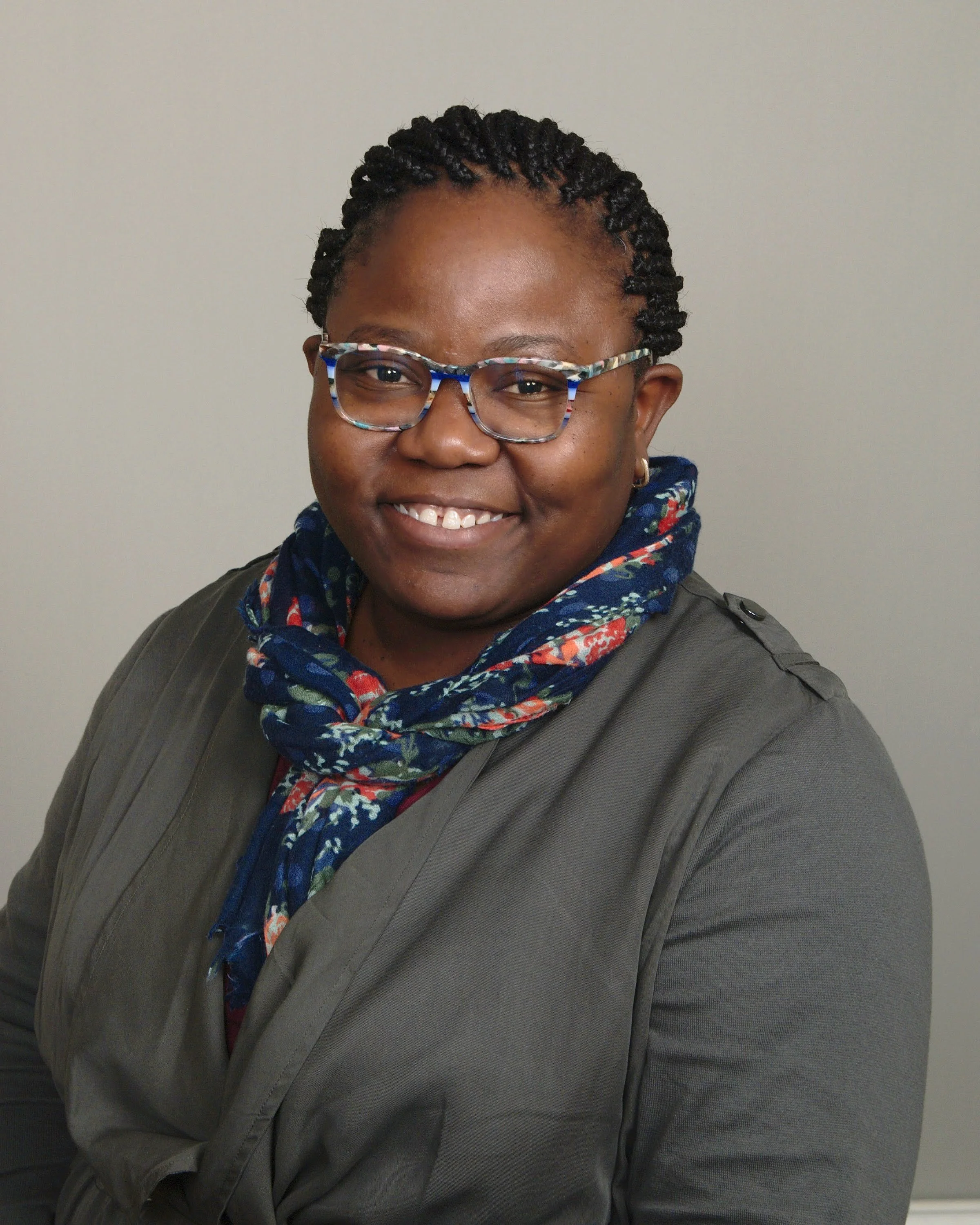 A portrait of a smiling woman with braided hair, wearing glasses, a gray jacket, and a colorful scarf, standing against a plain background.
