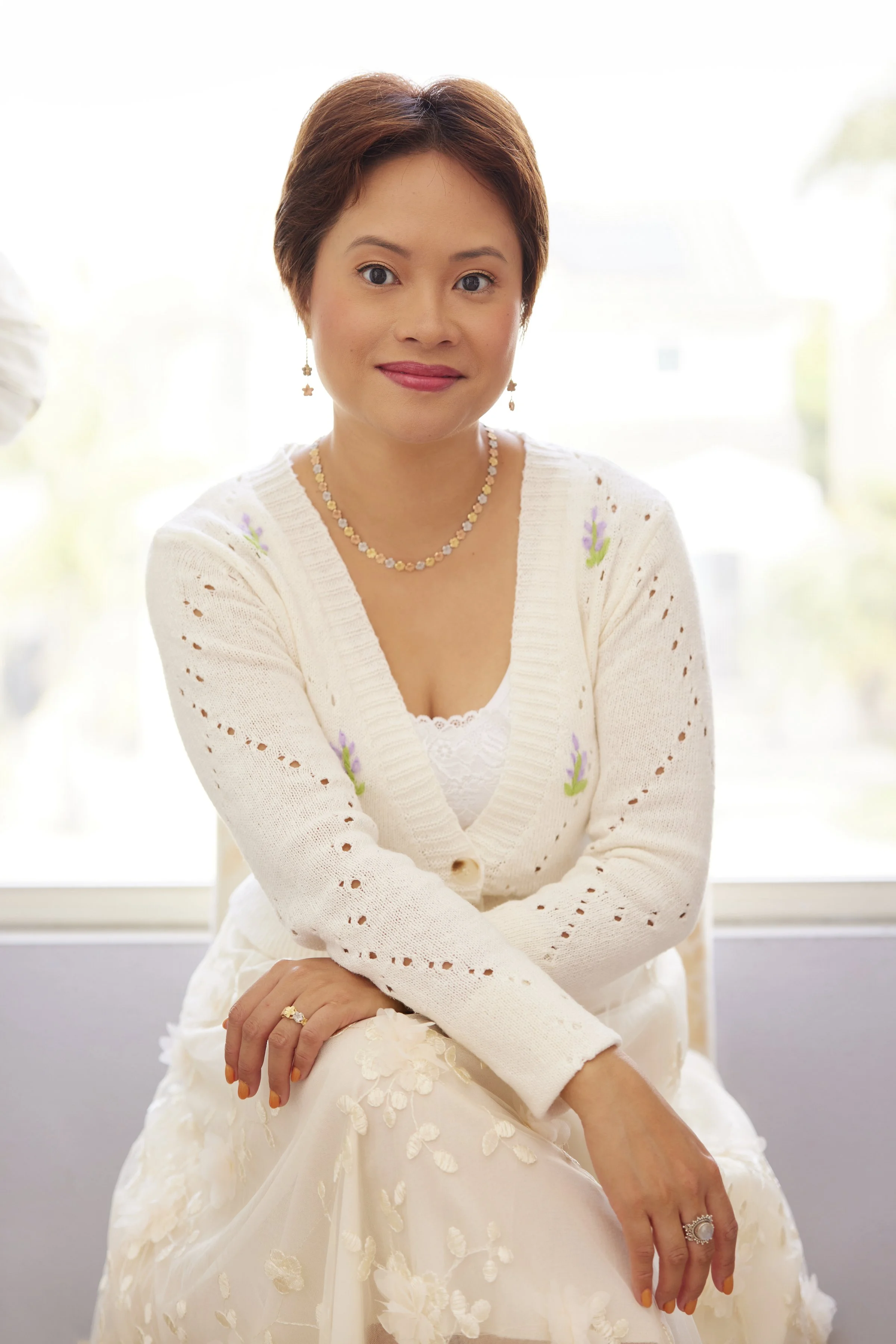 A woman with short brown hair and light skin, smiling, sitting near a window with a blurred outdoor background. She is wearing a white lace dress with floral details, a light-colored cardigan with embroidered flowers, and jewelry including a necklace, earrings, and rings.