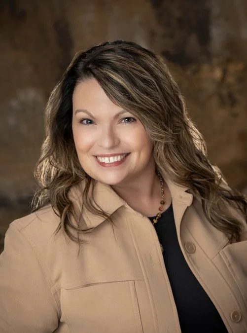 A woman with wavy brown hair and light skin smiling at the camera, wearing a beige jacket over a black top, with a blurred brown background.