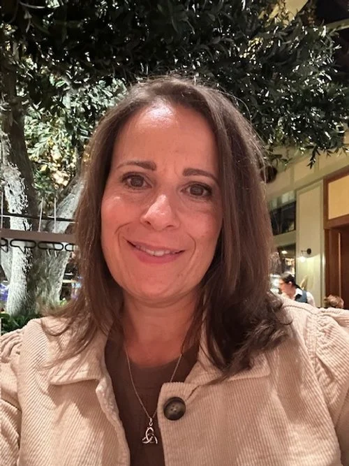 A woman smiling at a restaurant table with greenery and other people in the background.