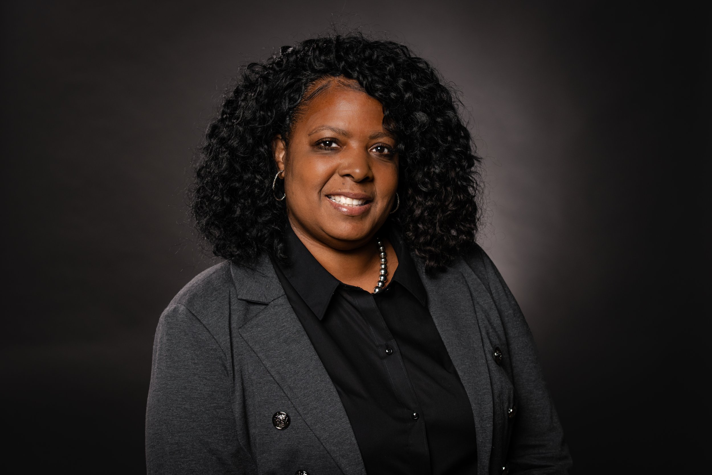Portrait of a woman with curly black hair, wearing a dark blazer and black shirt, and a pearl necklace, against a dark background.