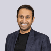 Headshot of a smiling man with black hair, wearing a dark blazer and black shirt against a light background.