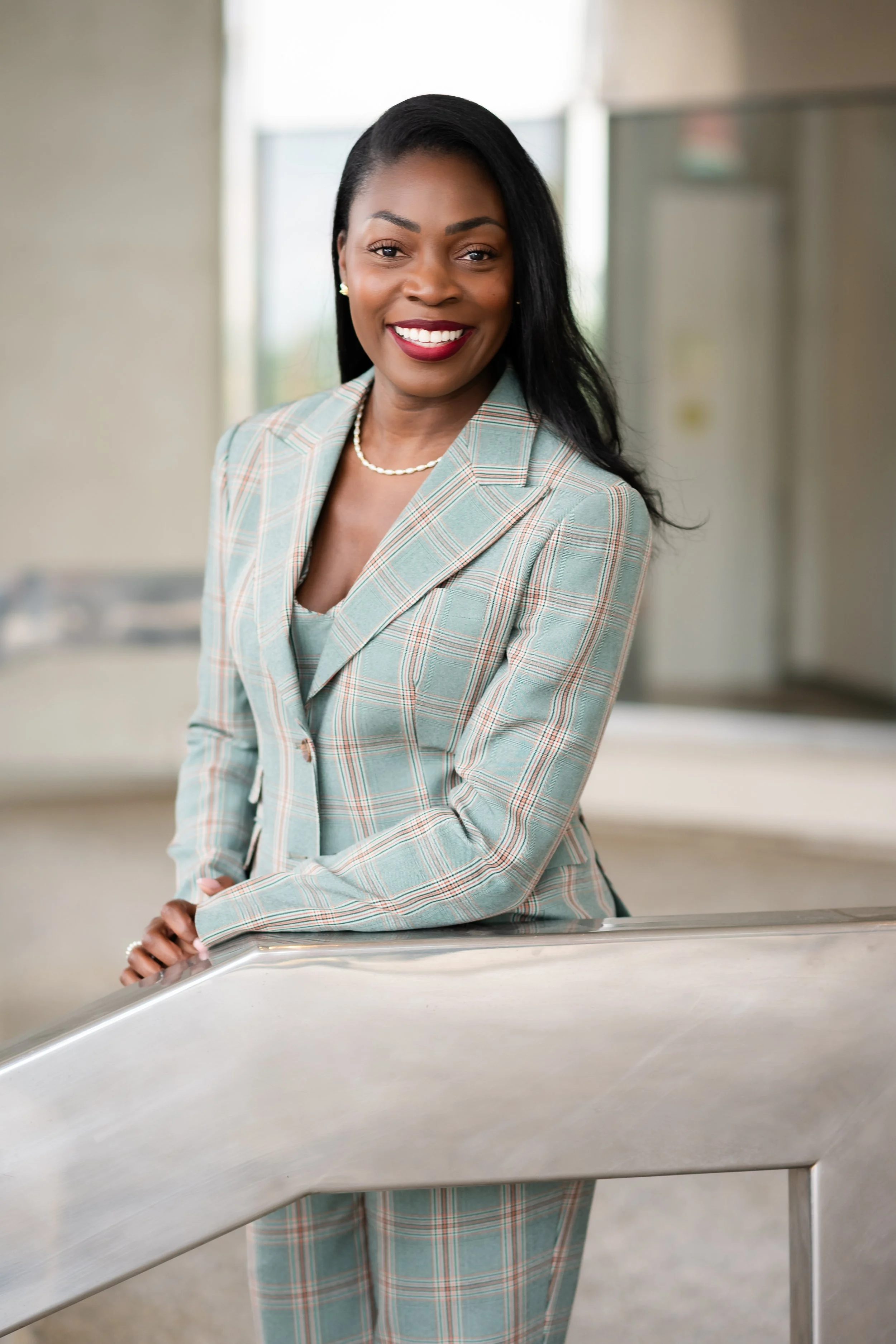 A woman with dark hair, wearing a light plaid suit and pearl jewelry, smiling and leaning on a metal railing in a professional setting.