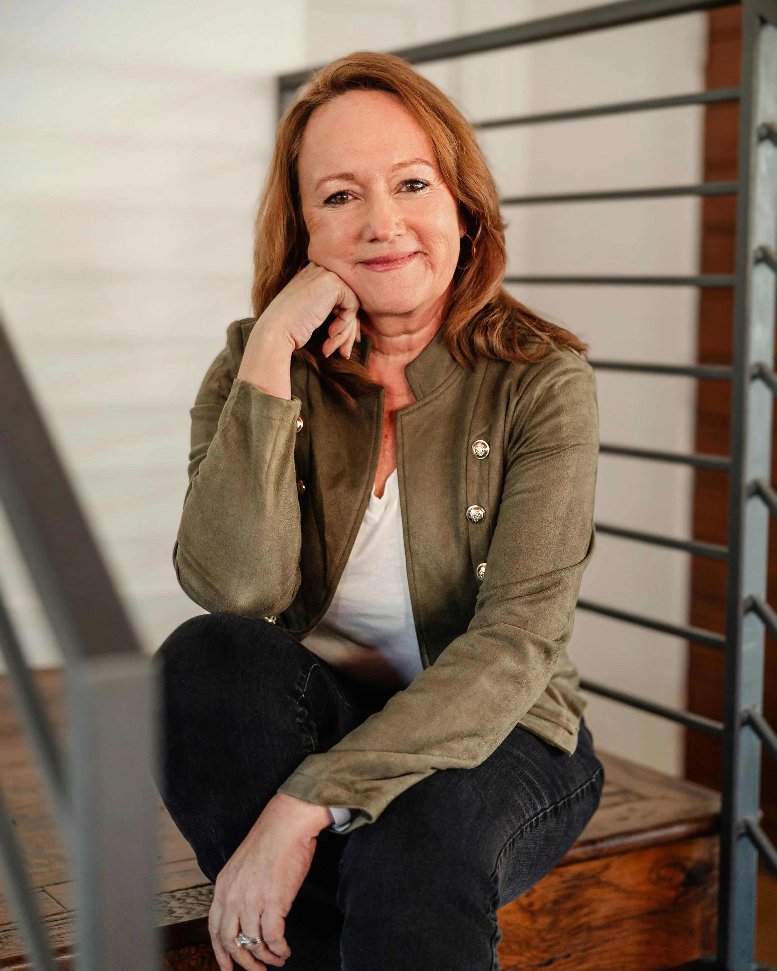A woman with shoulder-length red hair sitting on a wooden staircase, resting her chin on her hand, wearing a green jacket and dark jeans, smiling at the camera.