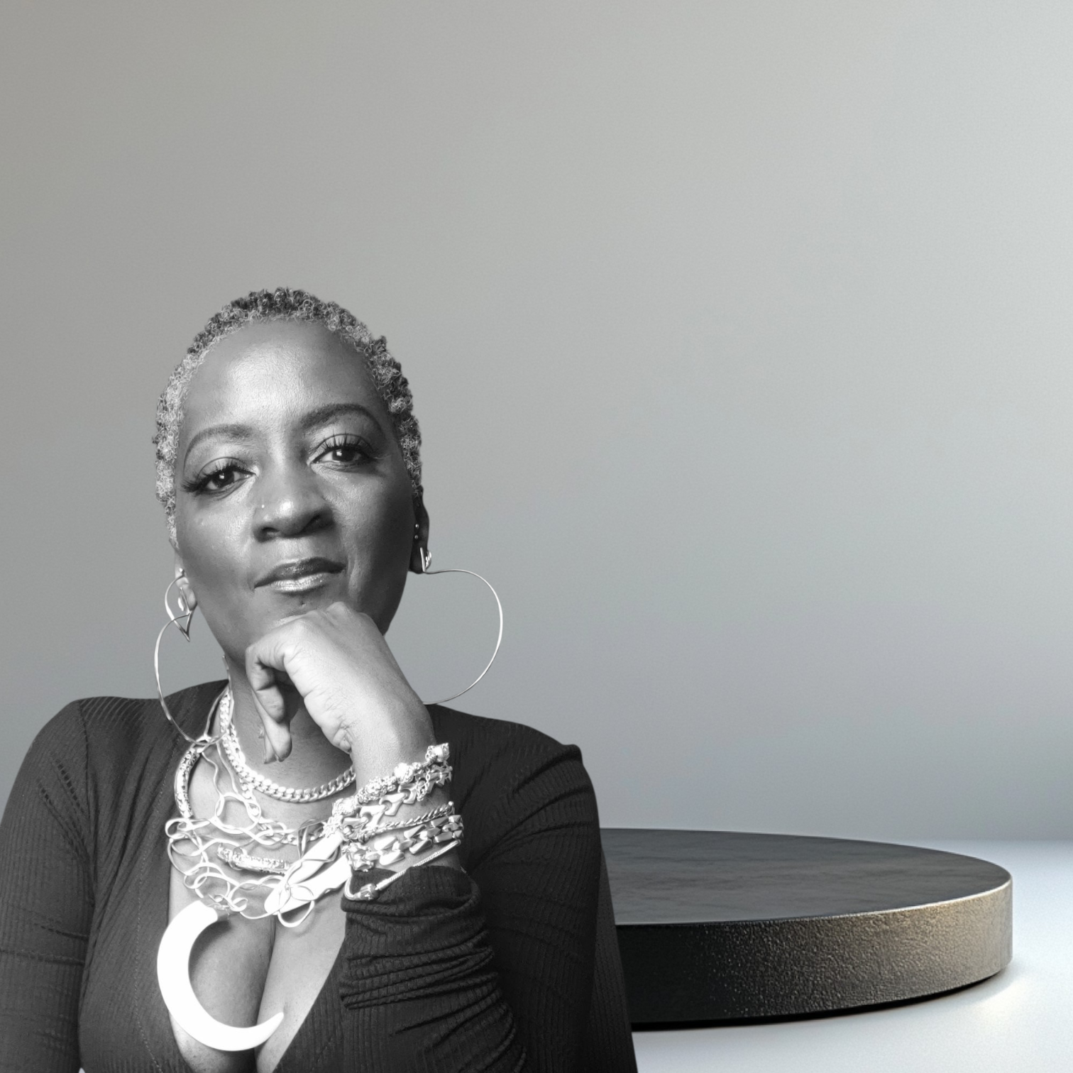A black-and-white portrait of a woman with short curly hair, wearing large hoop earrings and layered necklaces, seated with her hand resting on her chin, next to a round table.