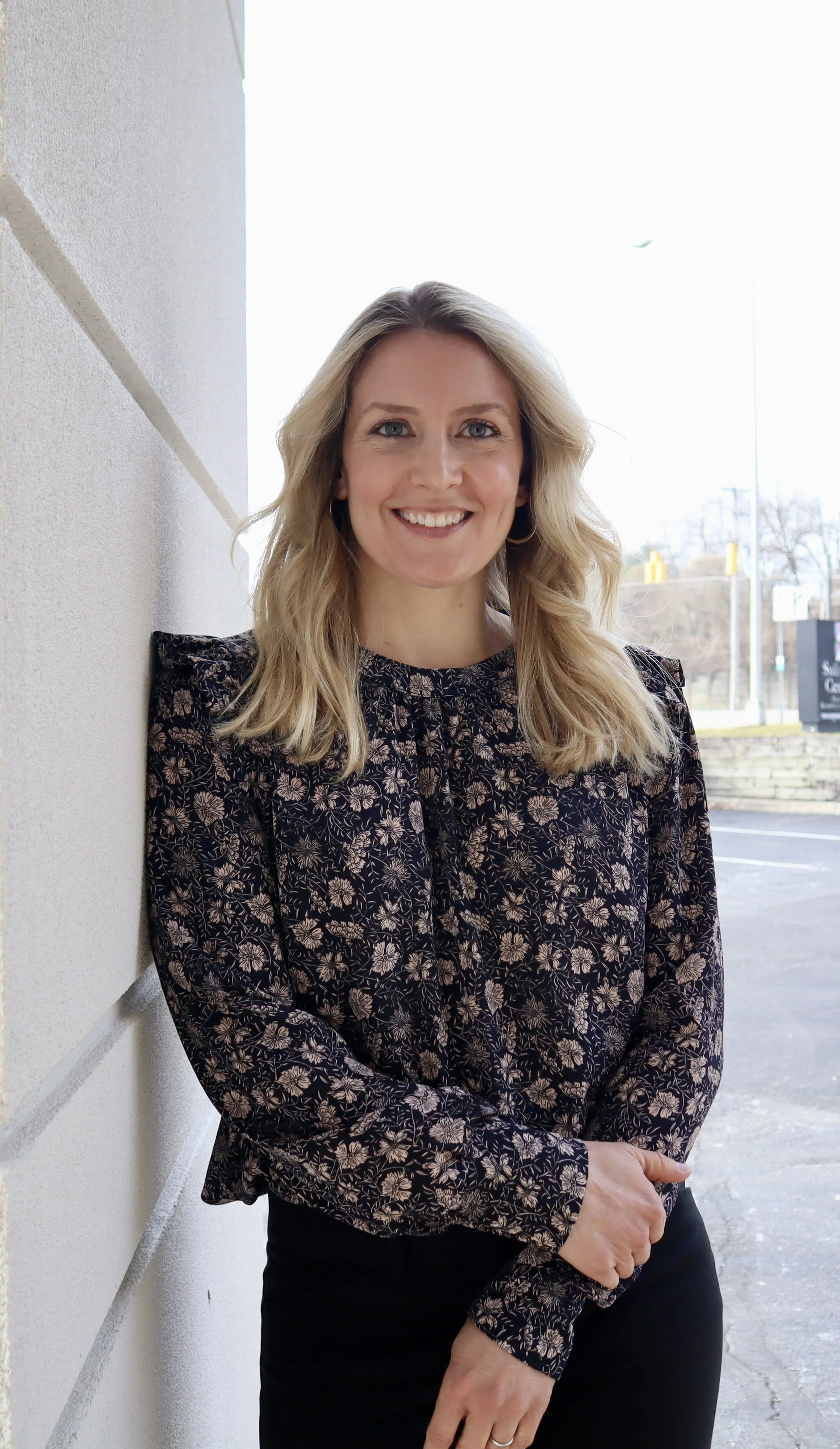 A woman with blonde hair, wearing a black long-sleeve floral blouse, stands outdoors against a white wall, smiling.