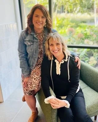 Two women smiling indoors, one sitting on a green couch and the other standing beside her, with a window and greenery outside in the background.