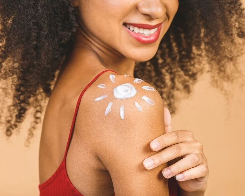 Woman with curly hair smiling, showing sunscreen in a sun design on her shoulder.